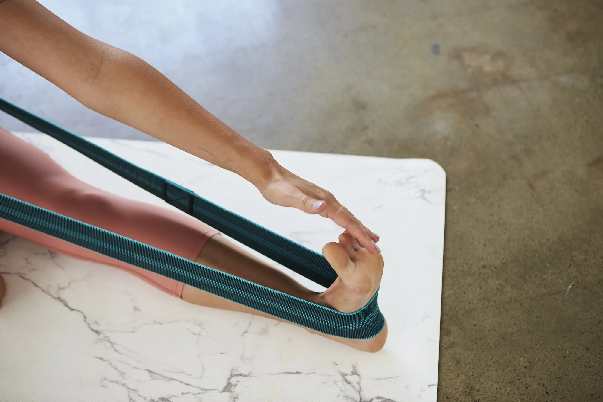 A person with bare arms practicing yoga, stretching their left arm and pointing their finger while sitting on a pink yoga mat. The scene is set on a marble surface with a concrete background.