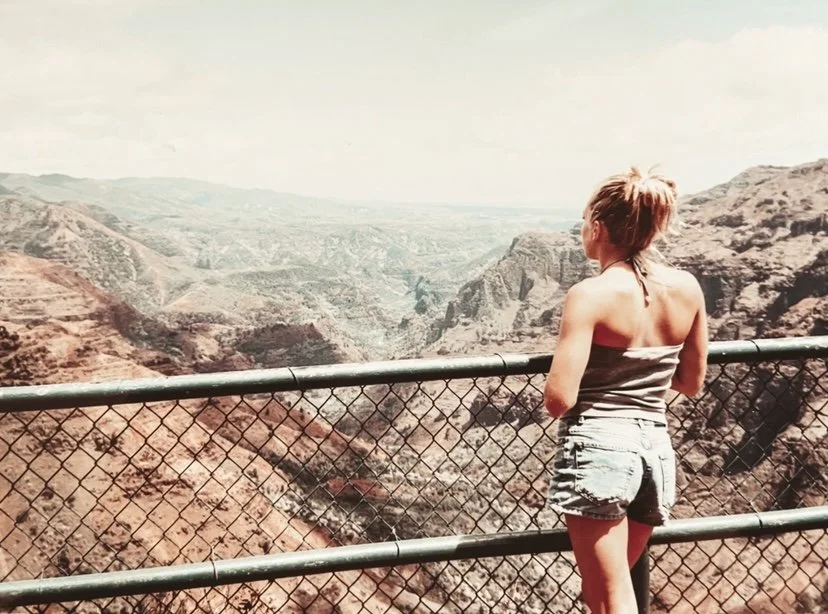 A woman stands at a lookout over the Grand Canyon, leaning on a metal fence and looking out at the vast canyon landscape.