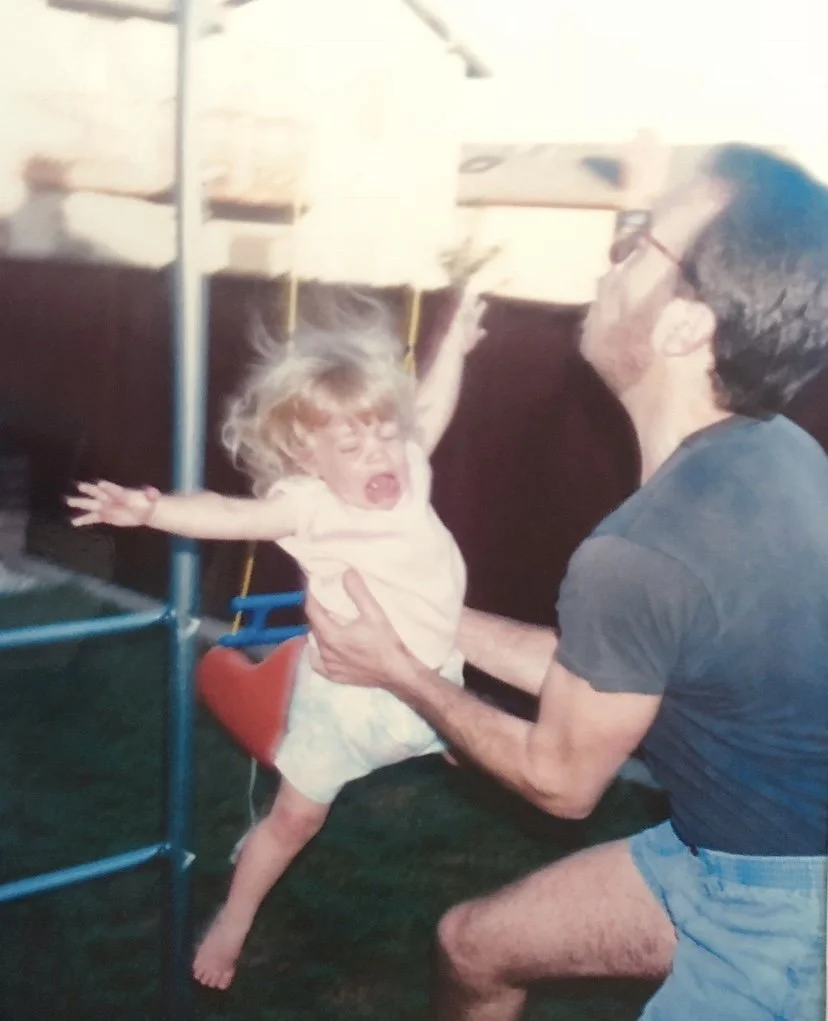 A young girl with curly blonde hair is being playfully swung by an adult man with glasses in an indoor setting, possibly a play area or home. The girl appears to be laughing or shouting, wearing a light-colored shirt and patterned shorts.
