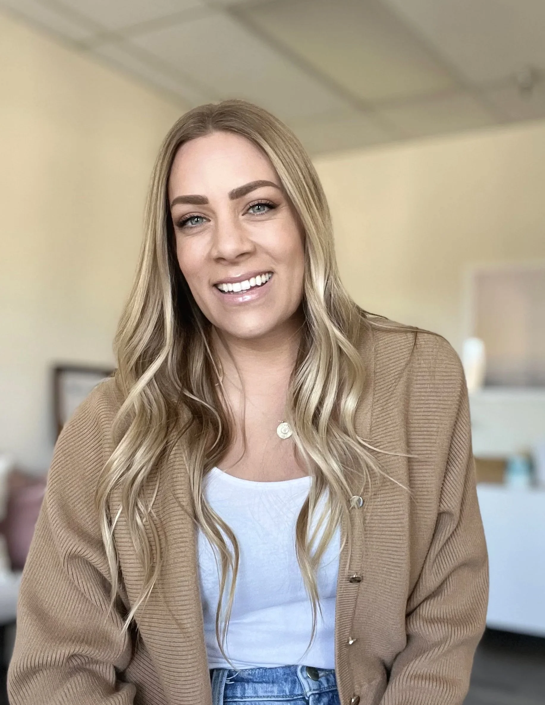 A smiling young woman with long wavy blonde hair, wearing a beige cardigan over a white top, standing indoors with a blurred background.