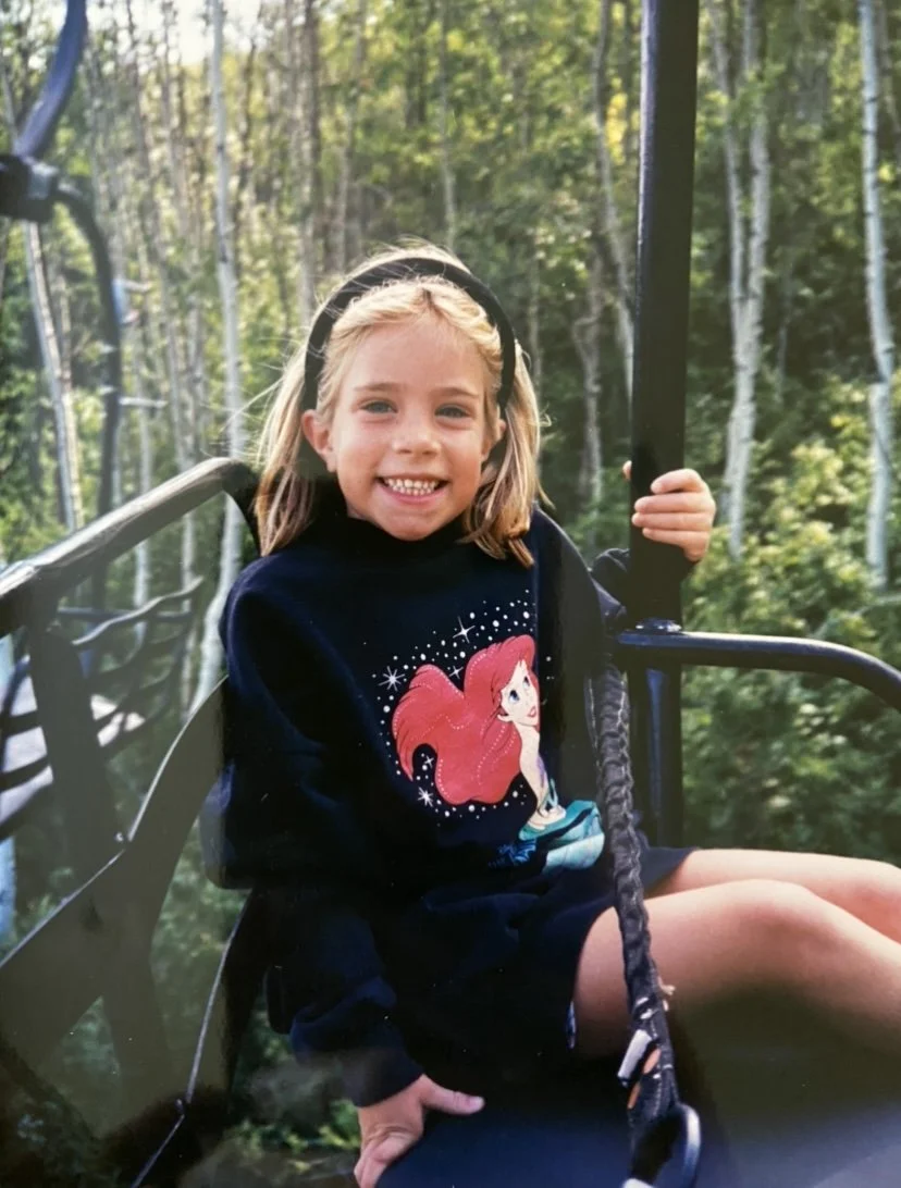 A young girl with blonde hair and a headband, smiling while sitting on a swing in a forested area.