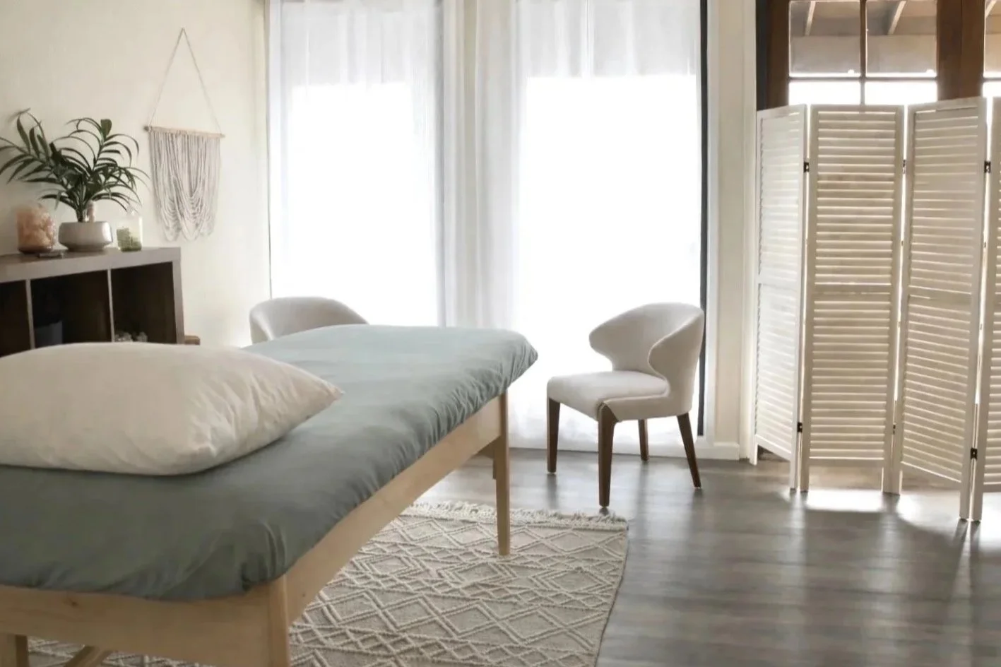 A bright room with a massage table, white pillow, white chair, decorative plants, a woven wall hanging, and a folding room divider, with sunlight coming through sheer curtains.
