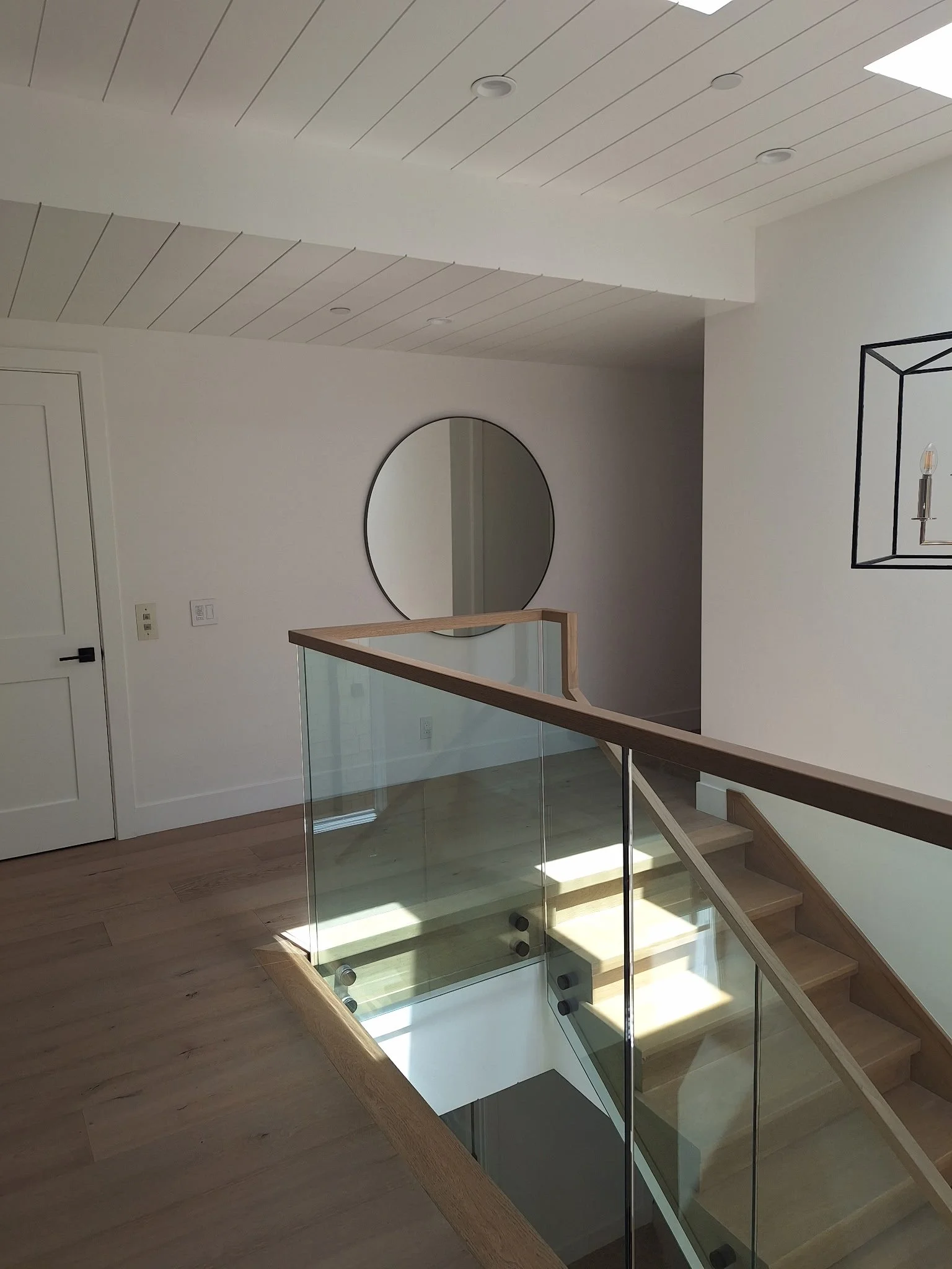 Interior of a modern house with a staircase and glass railing, white walls, a round mirror, and a wall sconce with a light bulb
