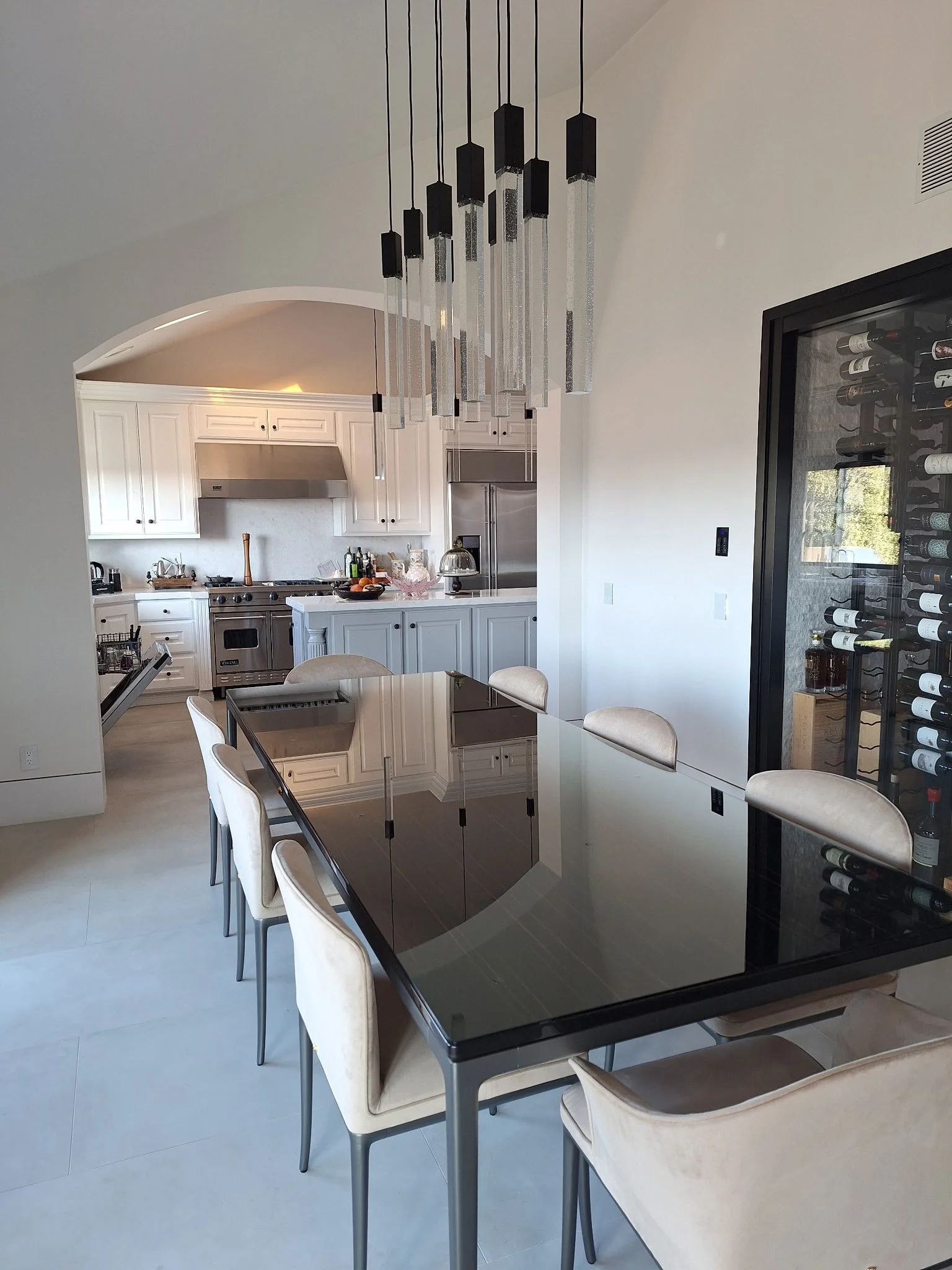 Modern kitchen and dining area with a black glass-top table, beige chairs, a wine refrigerator, and a chandelier.