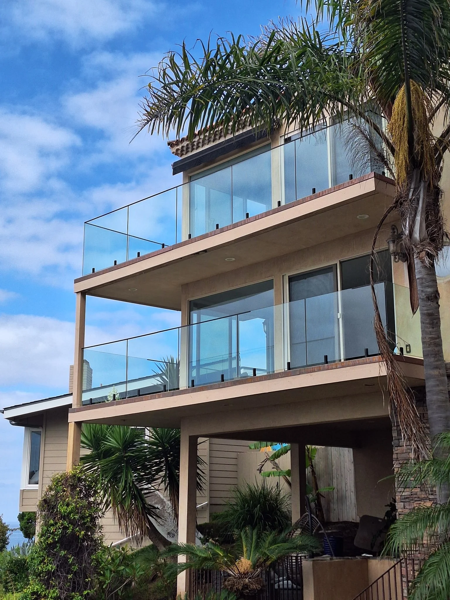 Multi-story modern house with glass balconies, palm trees, and lush greenery.