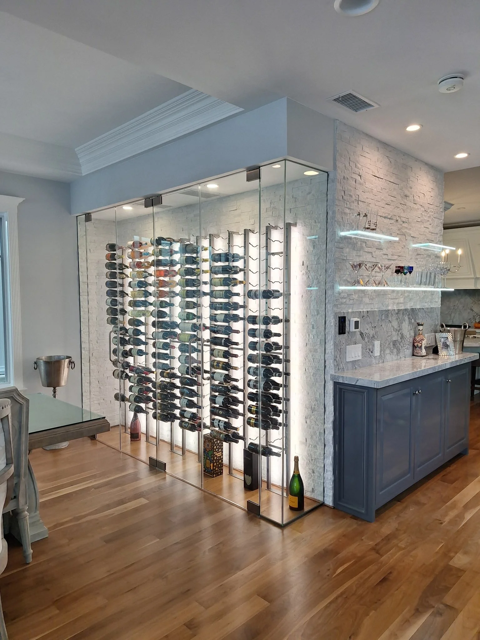 Glass-enclosed wine rack with multiple bottles of wine, located in a modern kitchen or dining area.