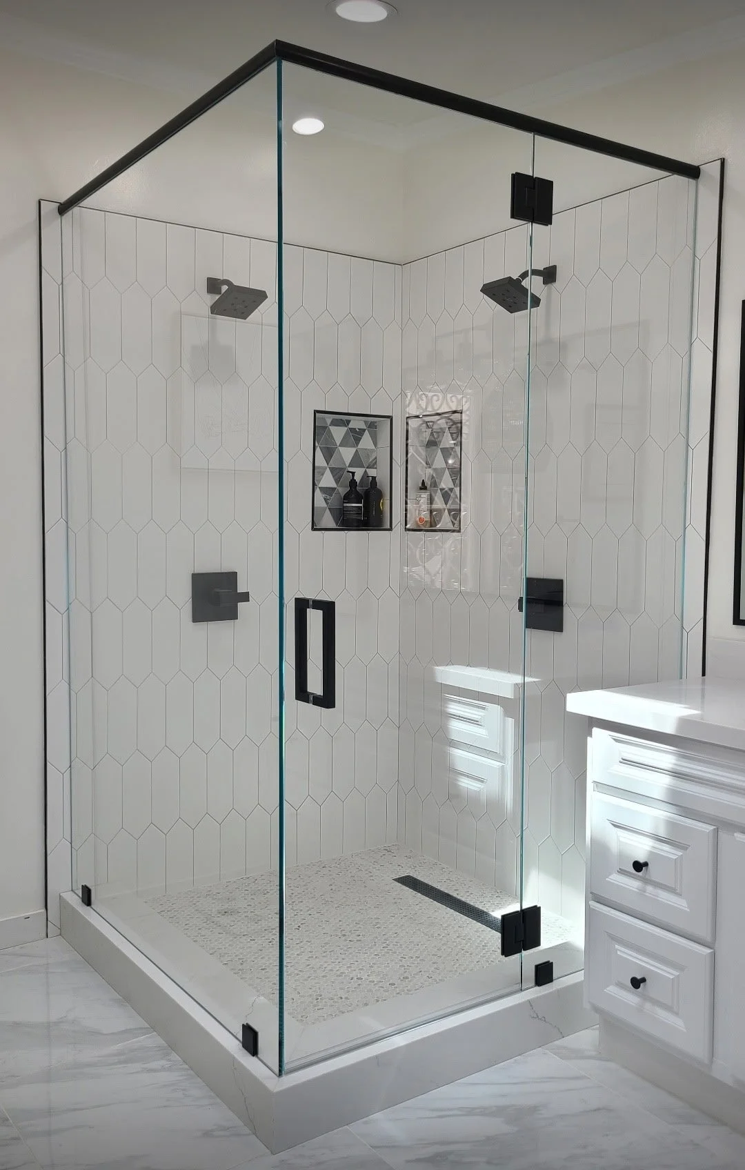 A modern glass shower enclosure with black fixtures in a bathroom with white hexagonal tiles on the walls and a white vanity visible to the right.