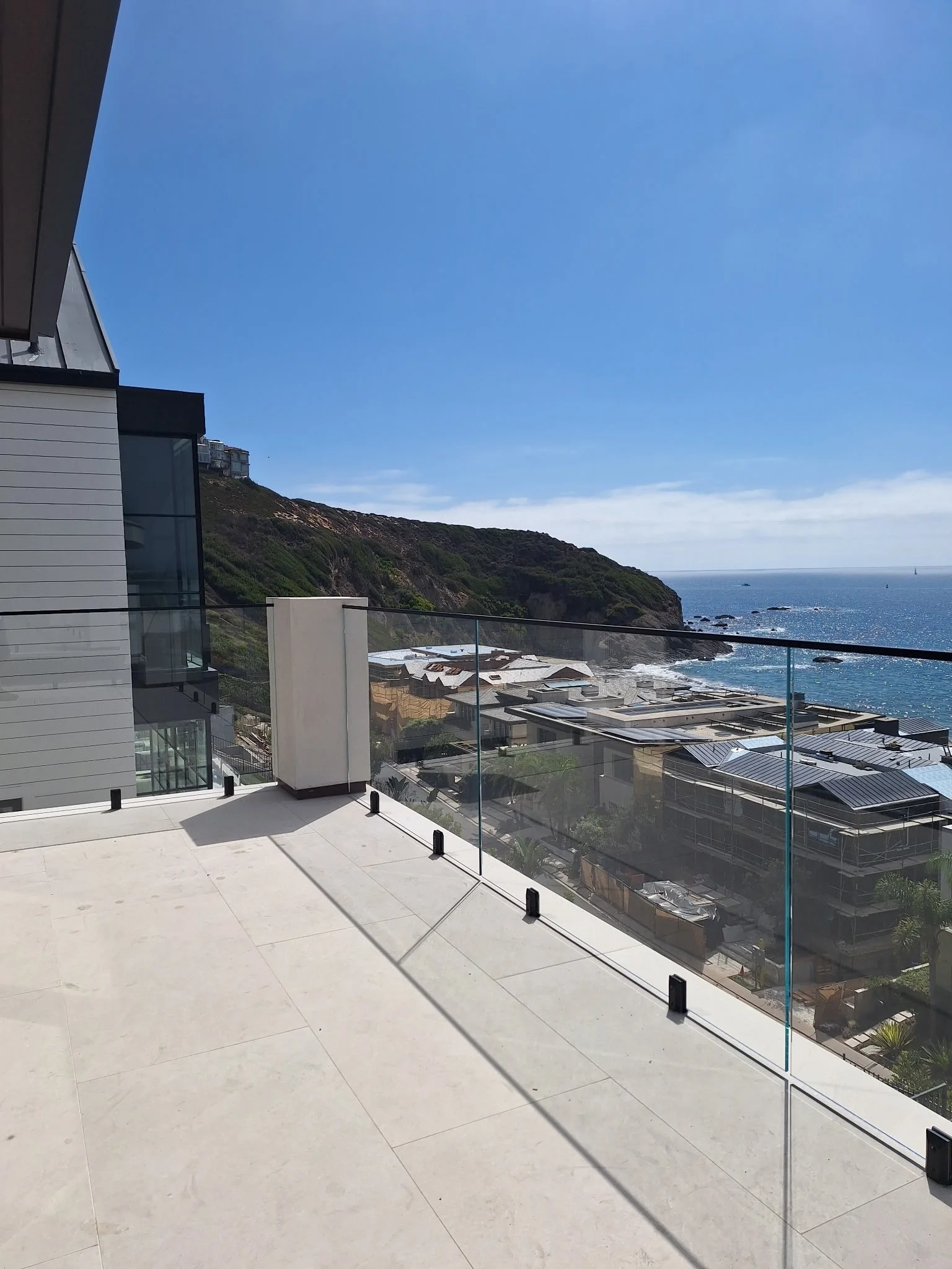 Balcony with glass railing overlooking a coastal landscape with buildings, green hills, and the ocean on a clear, sunny day.