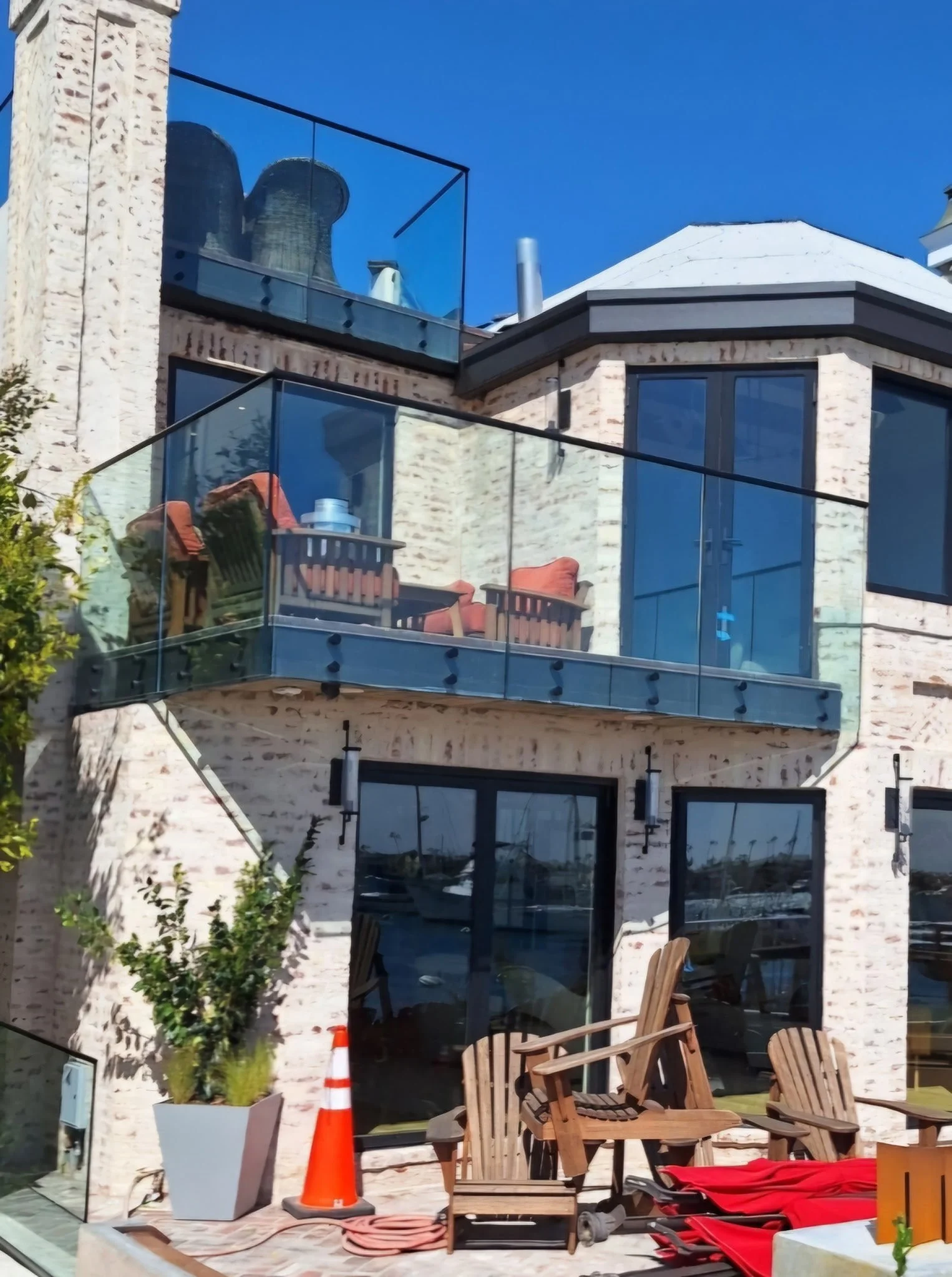 Modern multi-story building with large glass windows and glass balcony railings. The exterior is made of light-colored bricks, and there are outdoor chairs and some construction items on the ground level.