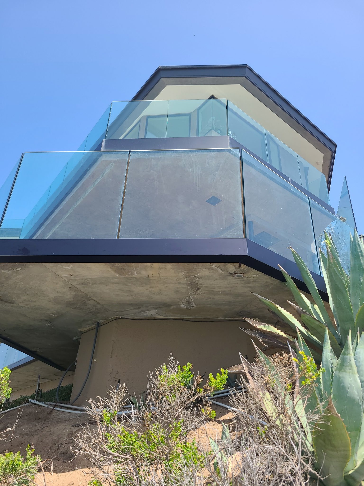 Low-angle view of a modern house with glass balcony and concrete structure, surrounded by desert plants and cactus under a clear blue sky.