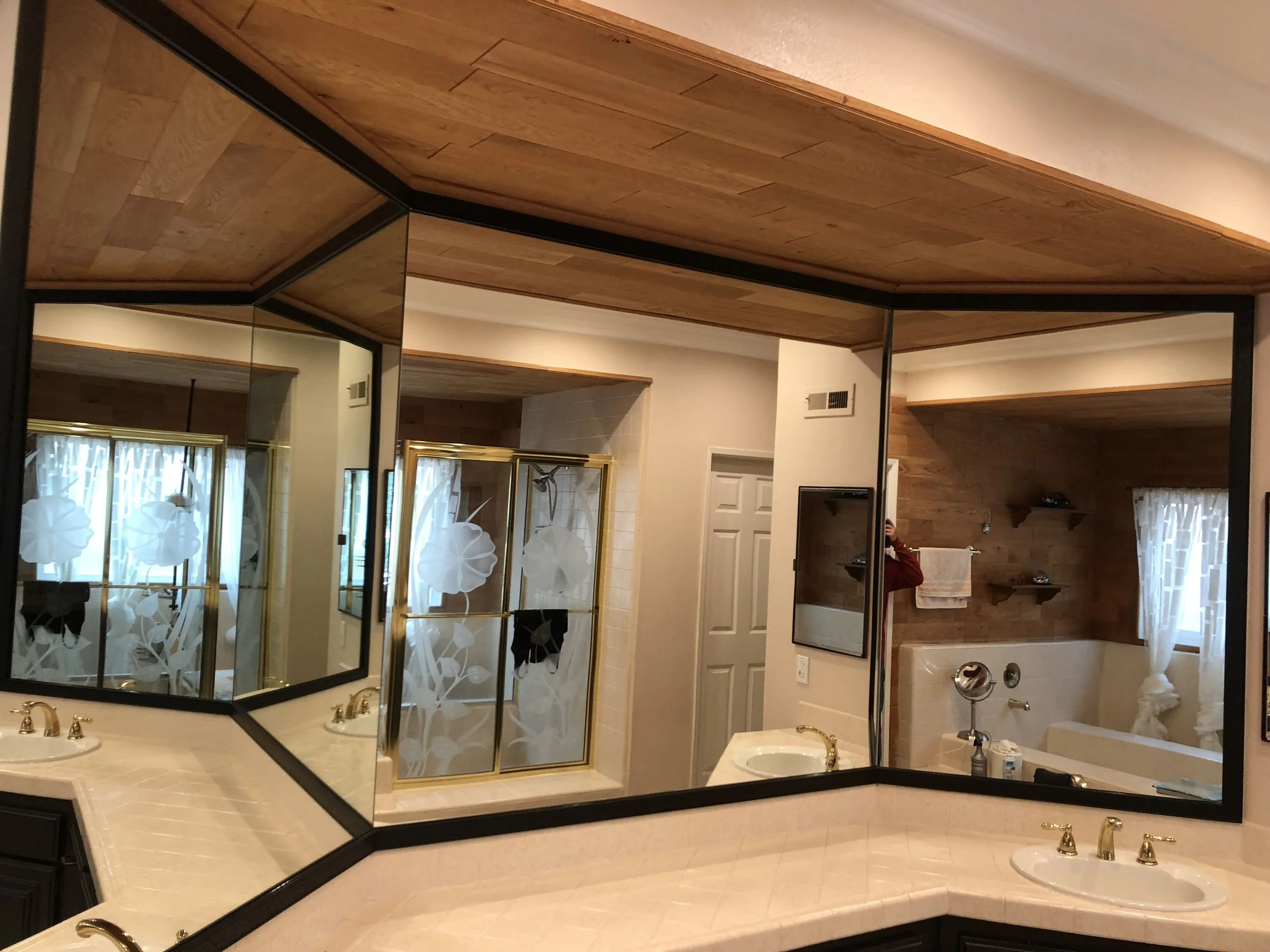 A bathroom with a large triangular mirror above a beige countertop with gold fixtures. There is a shower with a gold frame and frosted glass doors with floral patterns, a toilet, a bathtub, and wooden ceiling and walls. A window with white curtains is reflected in the mirror.