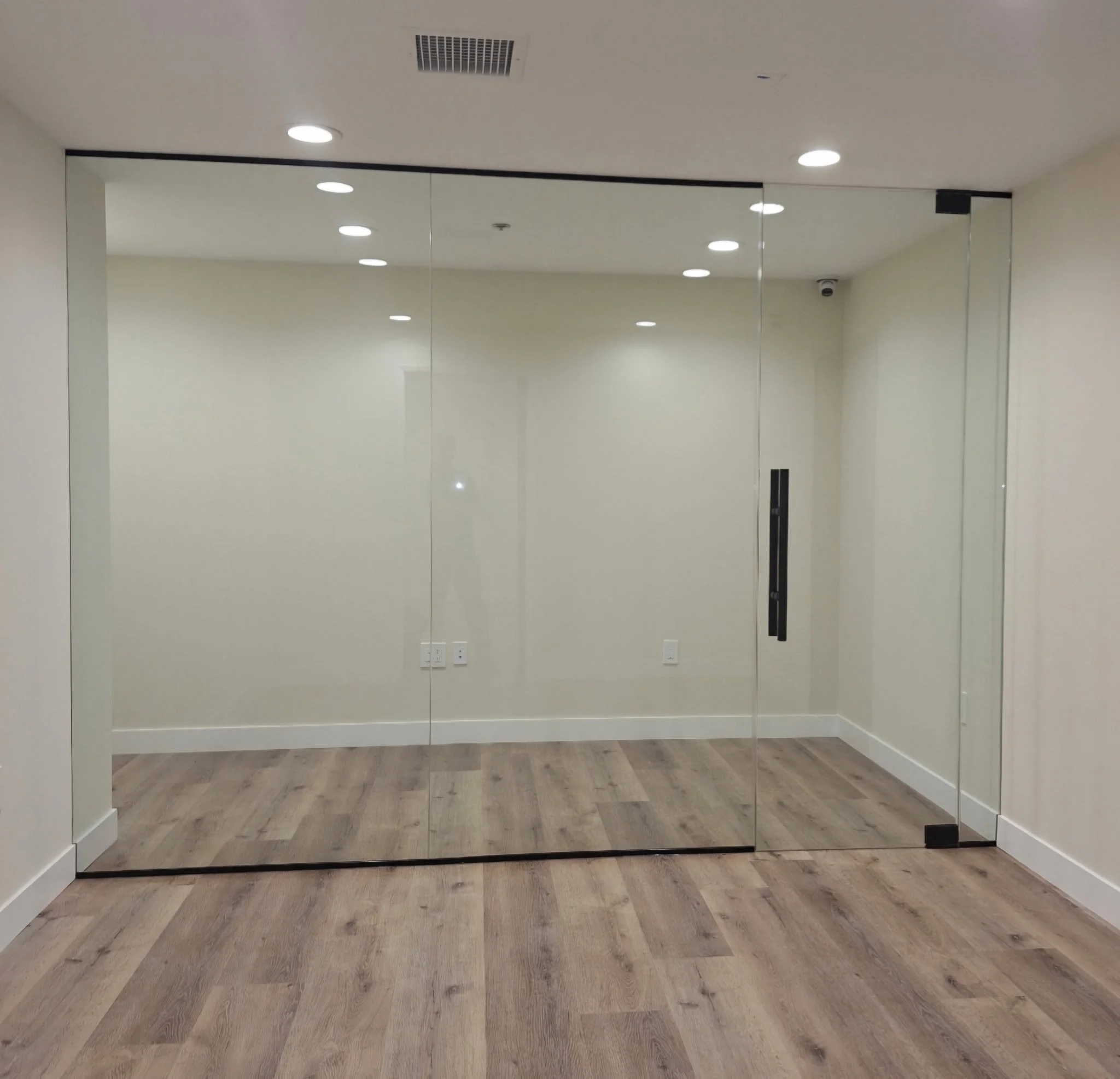 Empty glass-walled room with hardwood floors, white walls, ceiling lights, a black sliding door handle, and a security camera in the upper corner.