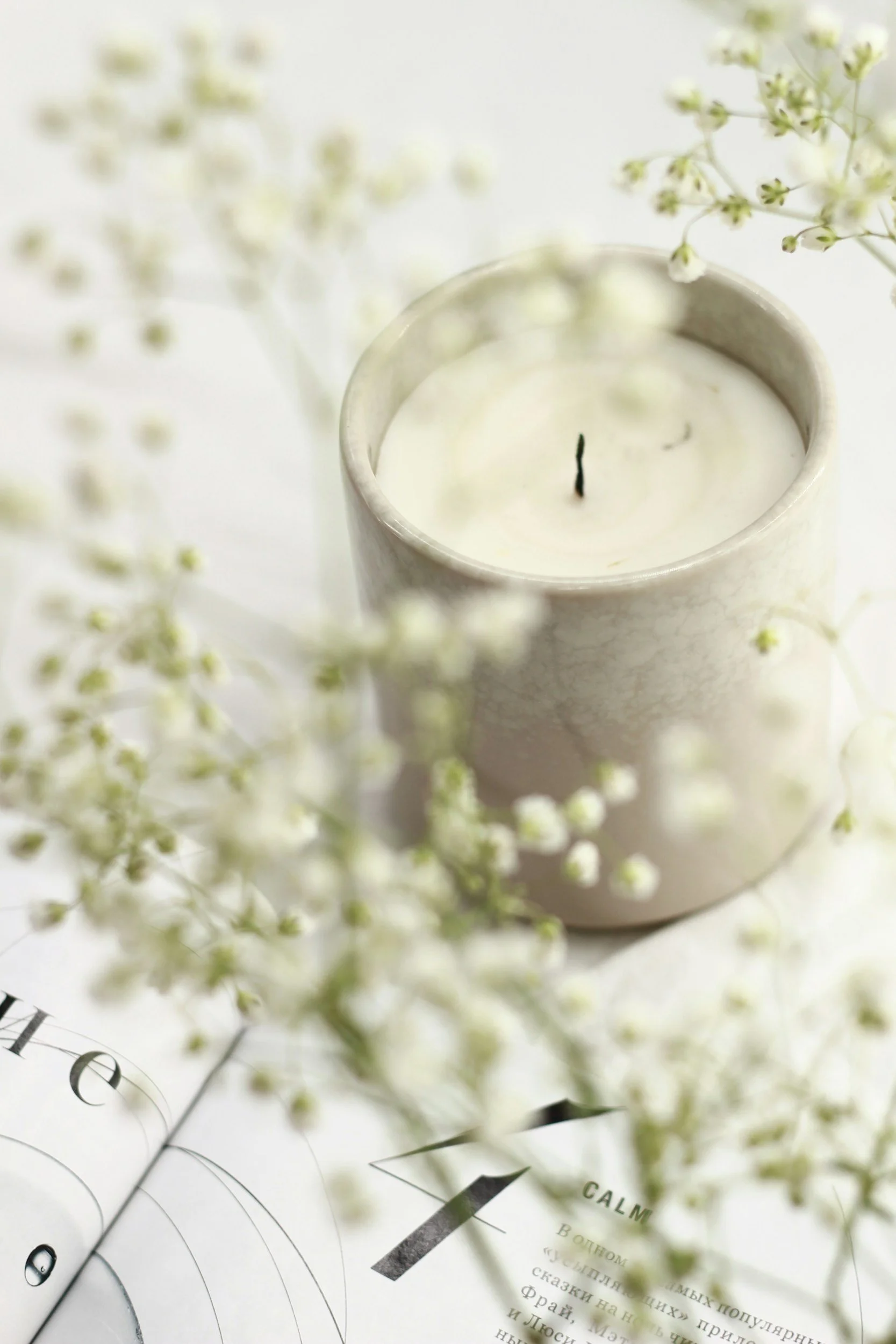 A white candle in a ceramic holder surrounded by small white flowers and an open book.