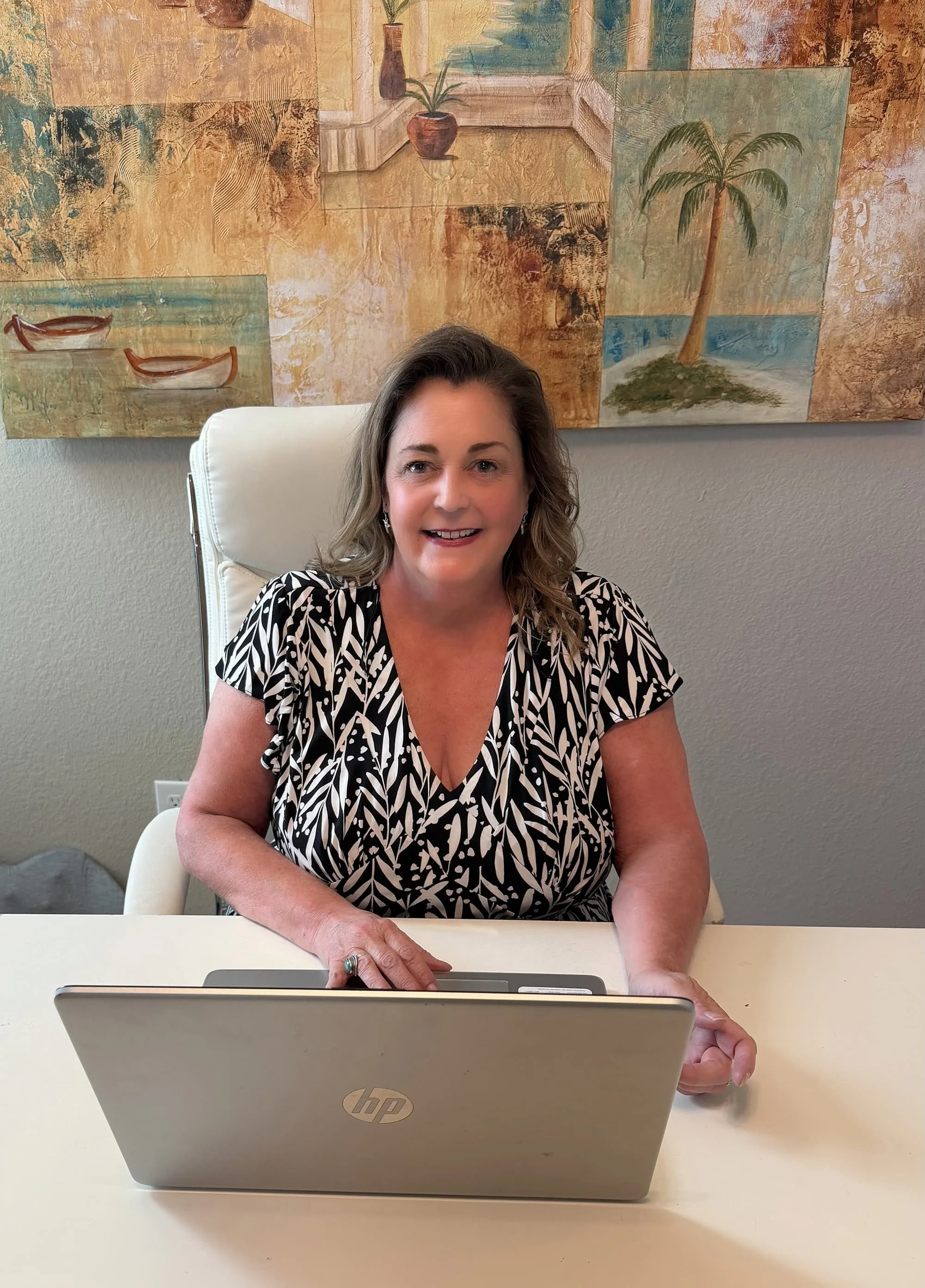 A woman with shoulder-length wavy hair sitting at a desk, smiling, in front of a laptop, with a colorful painting of boats, plants, and trees hanging on the wall behind her.