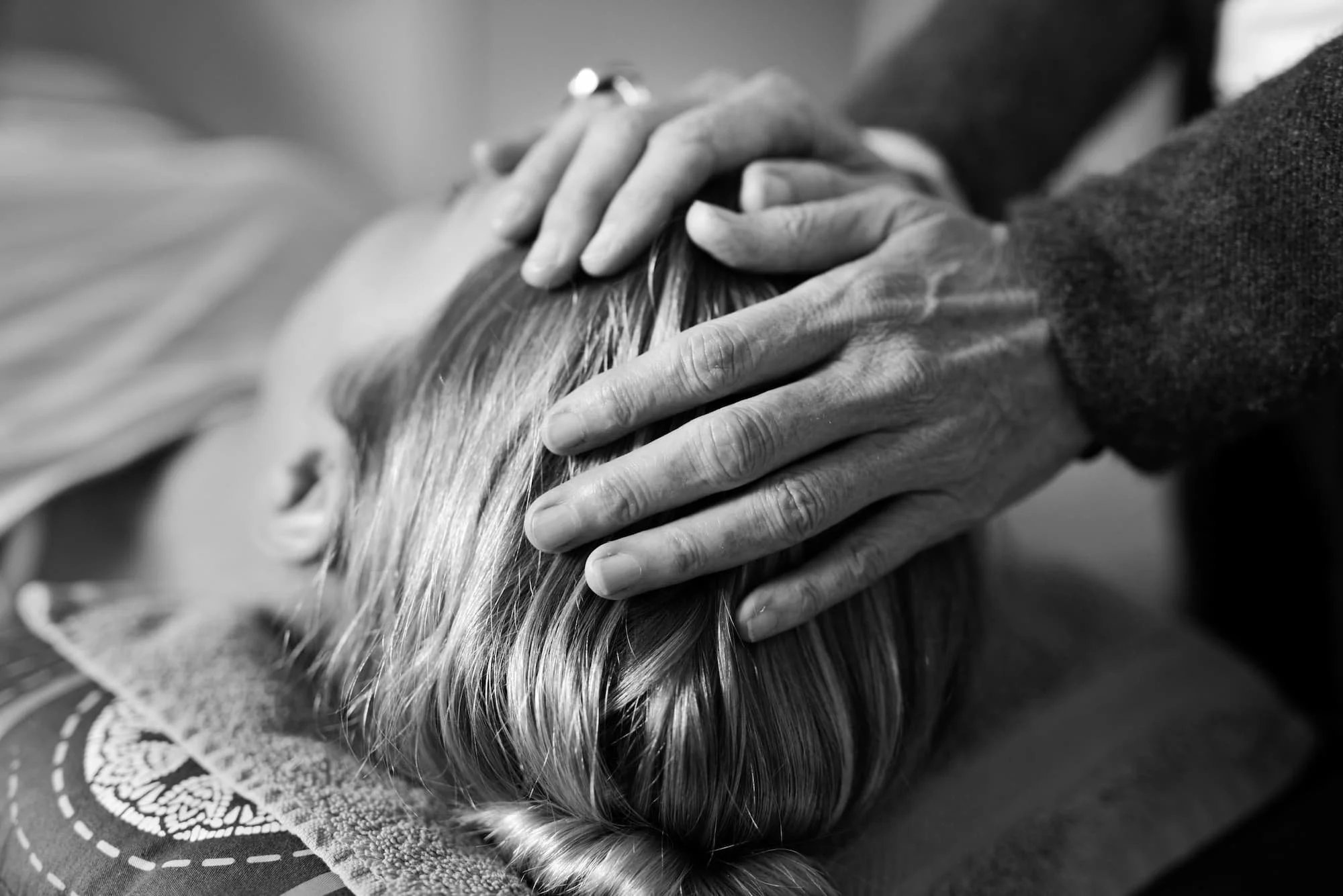 Black and white photo of hands gently holding the head of a person with long, blonde hair, lying face down on a textured surface.