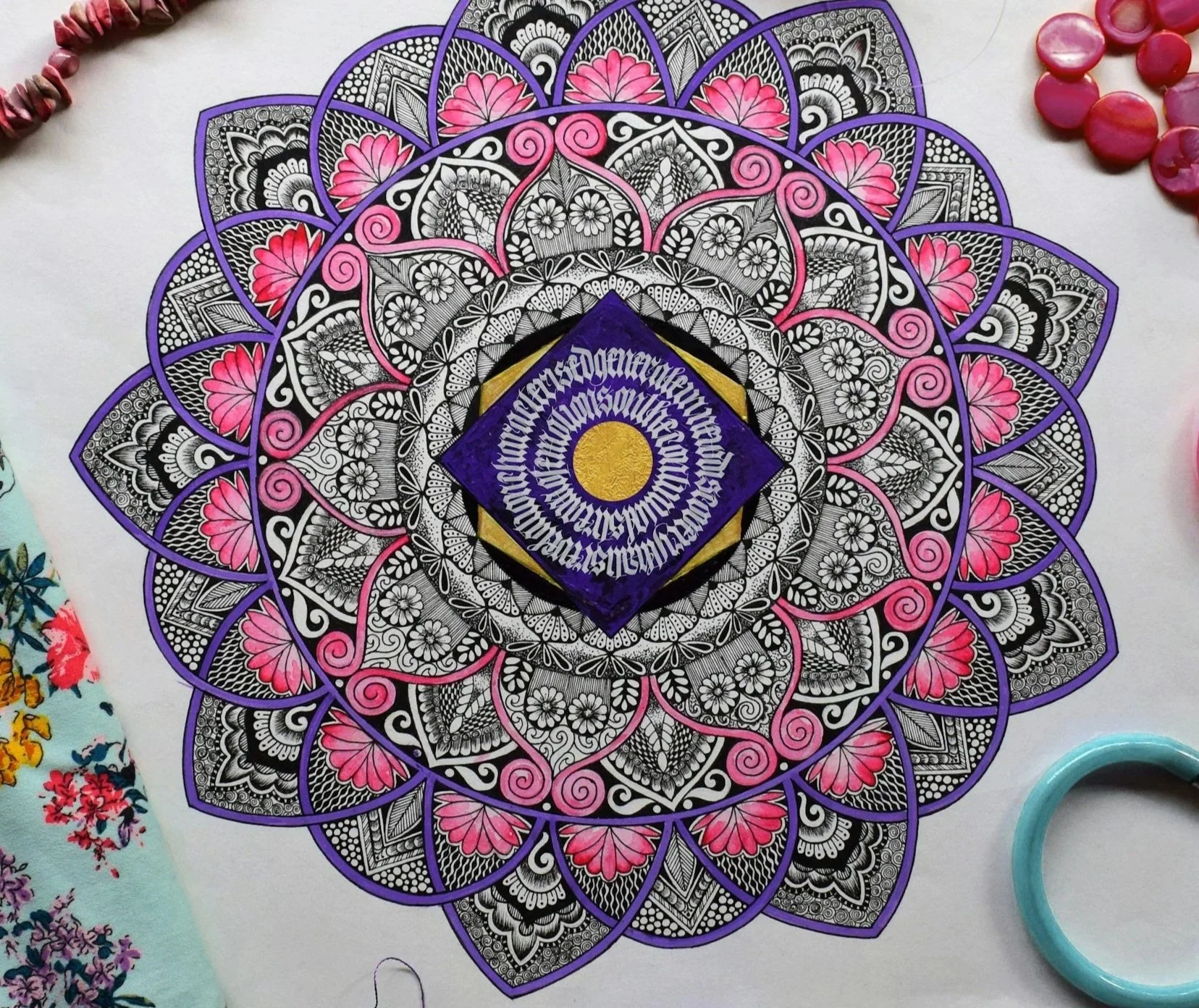 A detailed mandala drawing with black, white, pink, and purple colors. The central part has a purple diamond shape with intricate white text and a yellow circle. Surrounding it are various patterns in black, white, and pink, with some purple outlines, creating a symmetrical design.