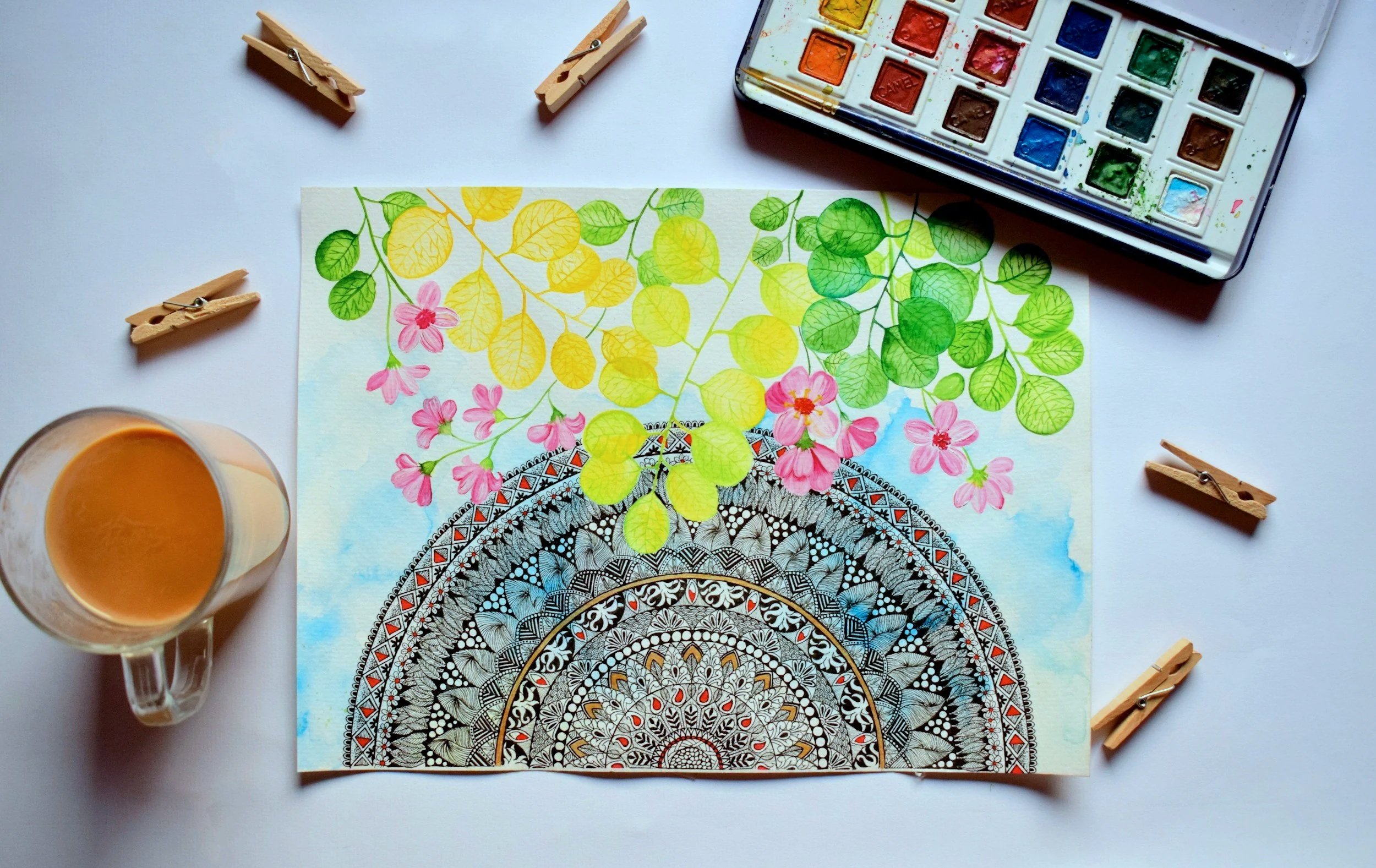 A watercolor and ink art piece of a colorful mandala pattern with green, yellow, and pink leaves and flowers, with watercolor paints, clothespins, and a glass of coffee on a white surface.