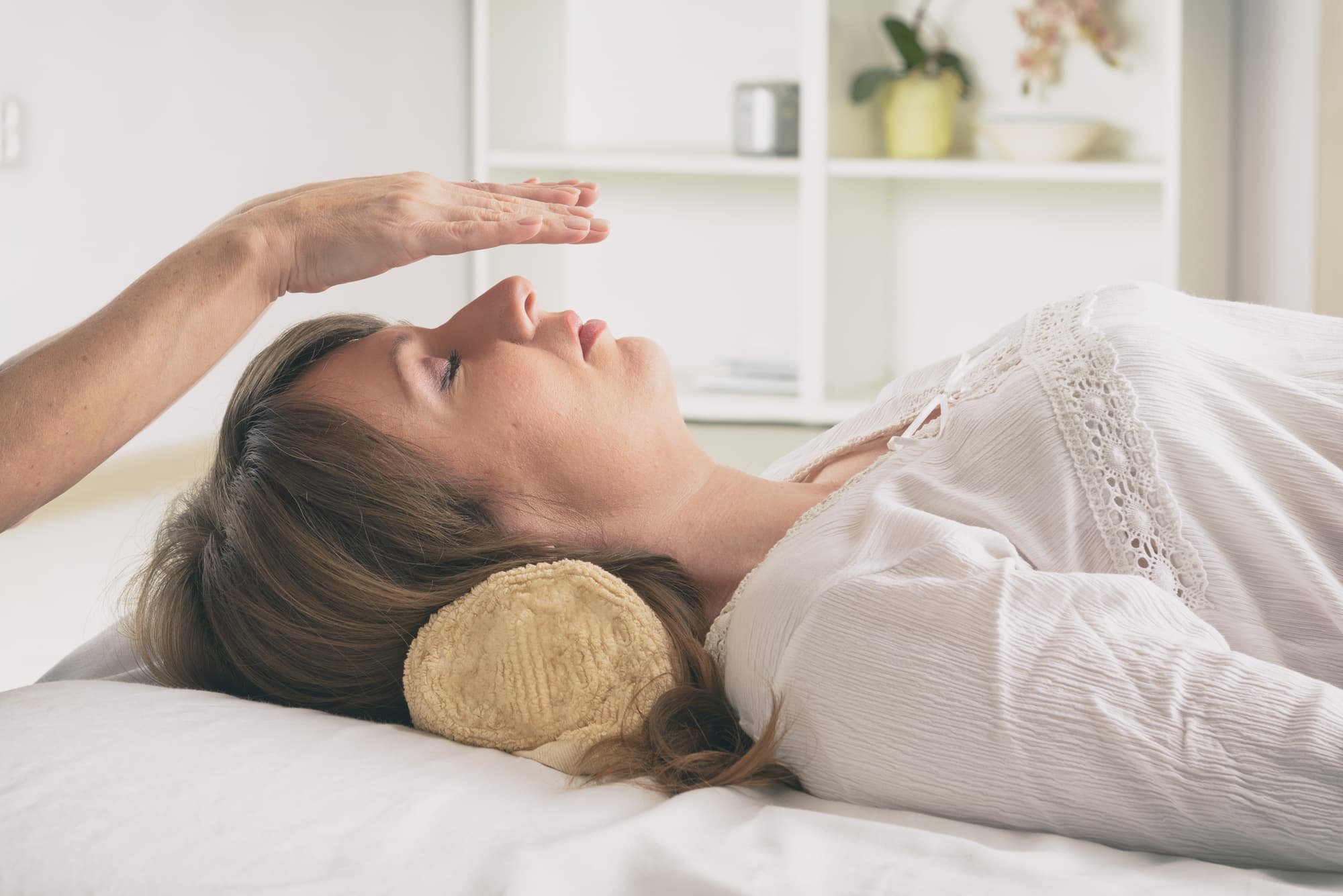 A woman receiving a forehead temperature check while lying on a bed with a pillow, in a bright room with shelves and plants in the background.
