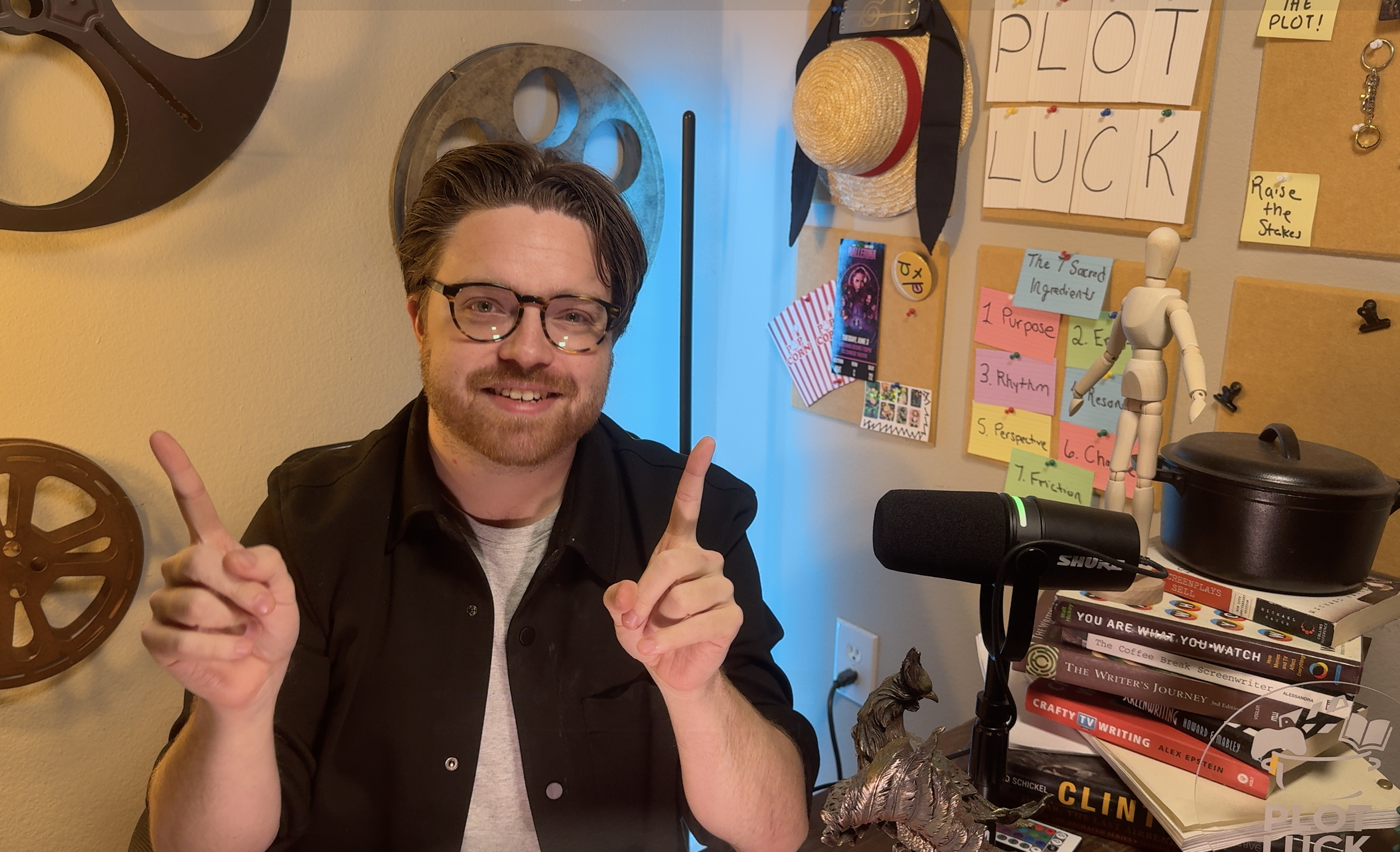A man with glasses and a beard smiling and pointing upward, sitting at a cluttered desk with books, a camera, and art supplies, in a room decorated with a corkboard, hats, and posters.