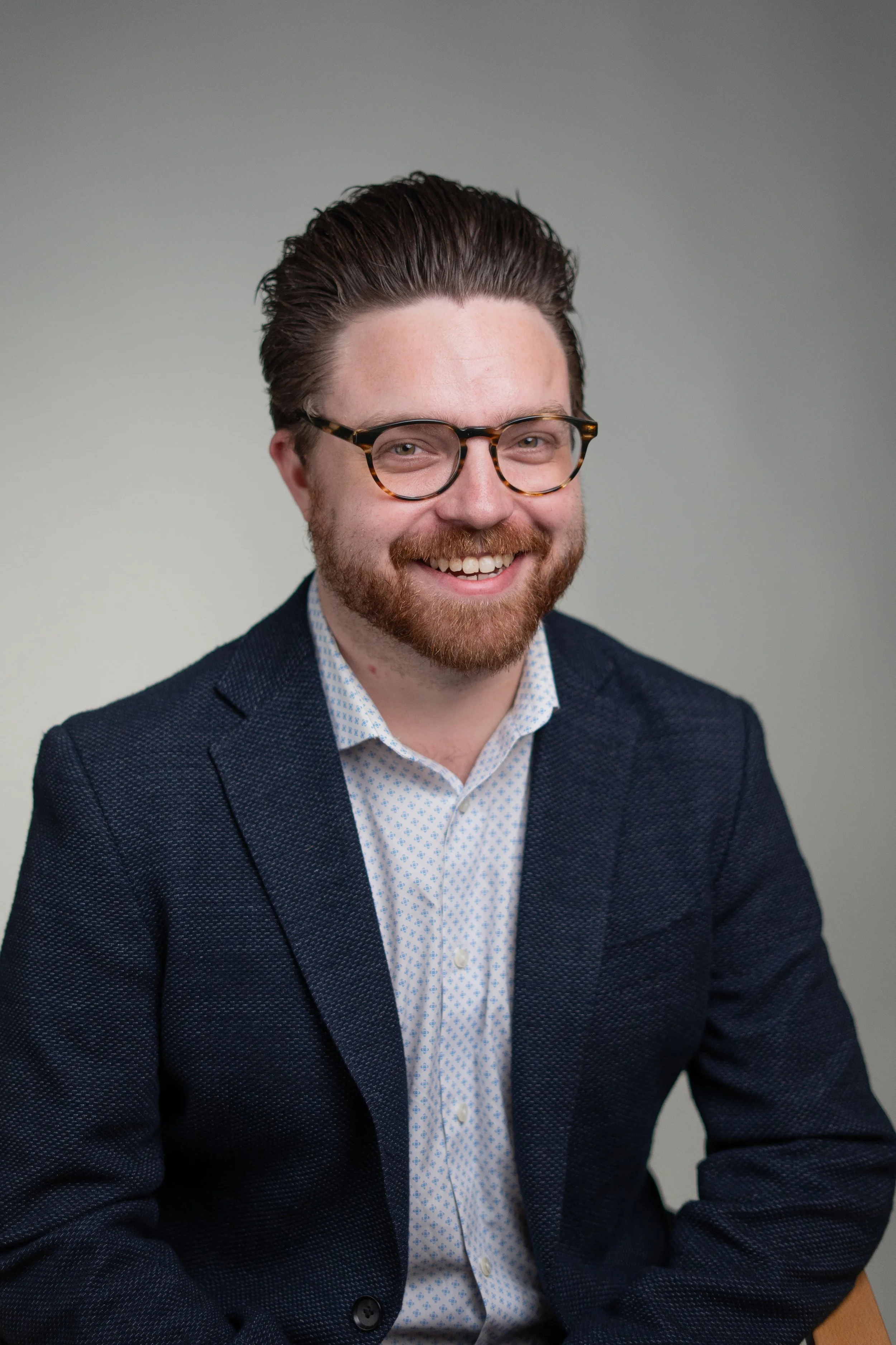 Headshot of a smiling man with brown hair, a beard, and glasses, wearing a dark blazer and a patterned shirt against a neutral background.
