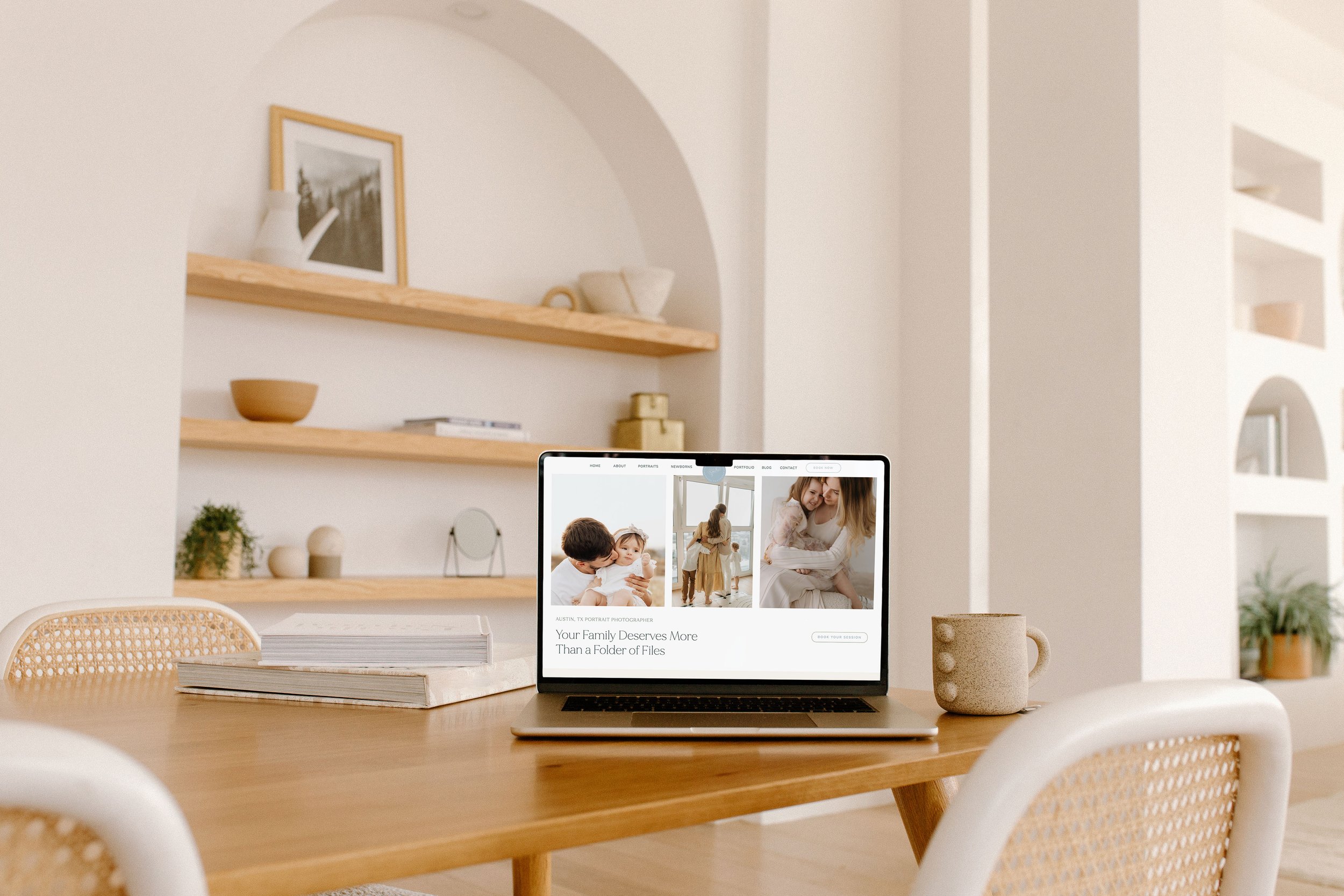 A workspace with a wooden table, a computer displaying a family portrait website, a white ceramic mug, and a stack of paper on the table. The background features white shelves with decorative items and a framed photo on the wall.