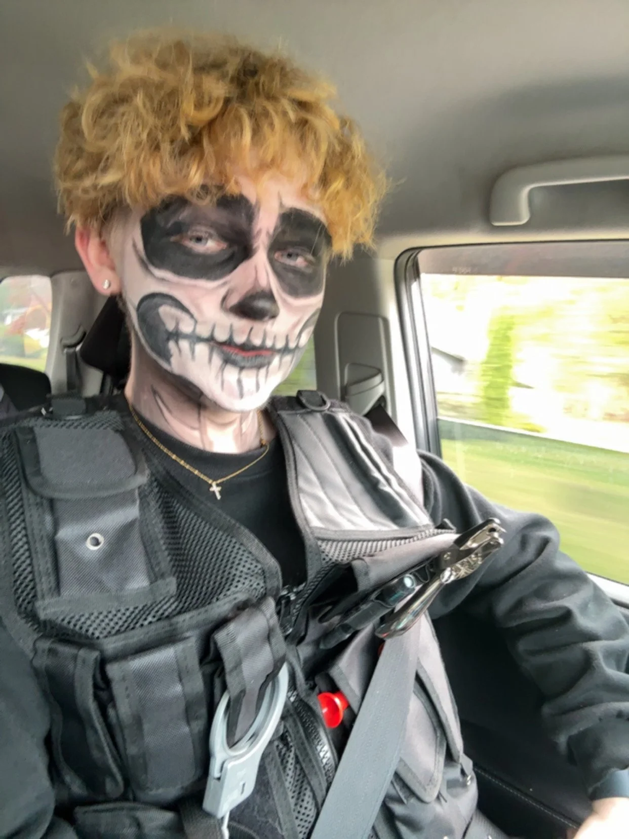 Person with skeleton face paint in the backseat of a vehicle, wearing tactical gear and holding a phone.