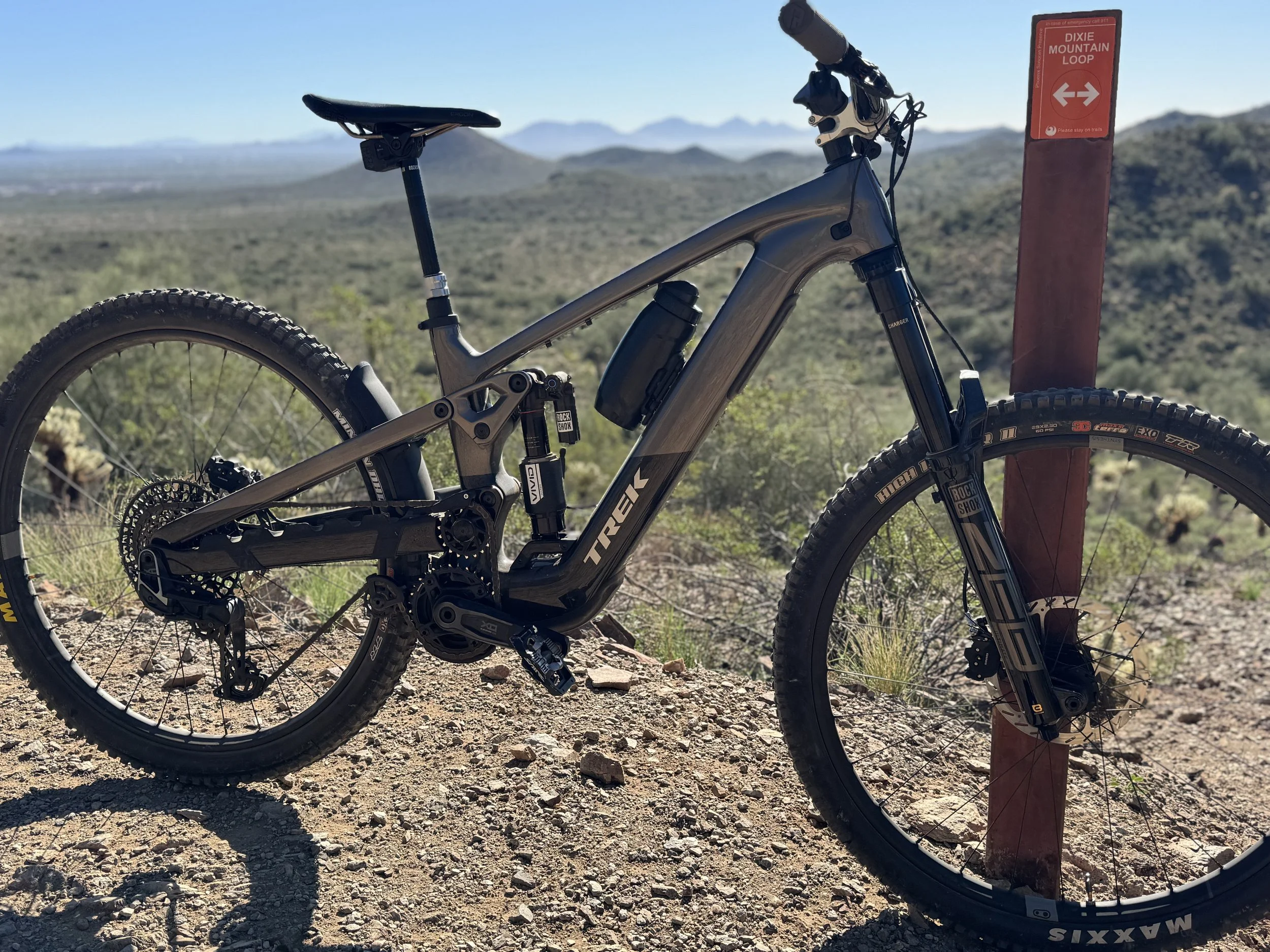 A Trek Slash+ 9.9 electric mountain bike resting against a trail marker pole in a desert landscape with distant mountains.