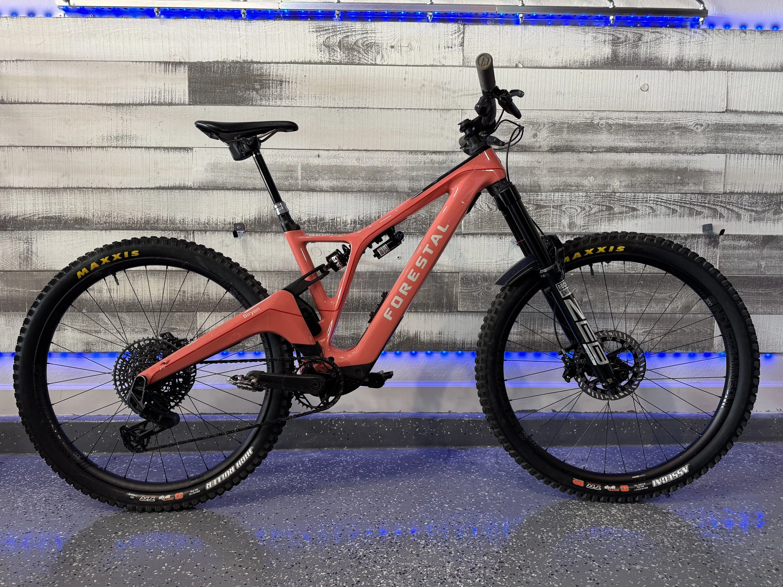 A pink Forestal Siryon mountain bike with black suspension forks and tires, displayed against a wooden wall with blue LED lighting and a speckled gray floor.