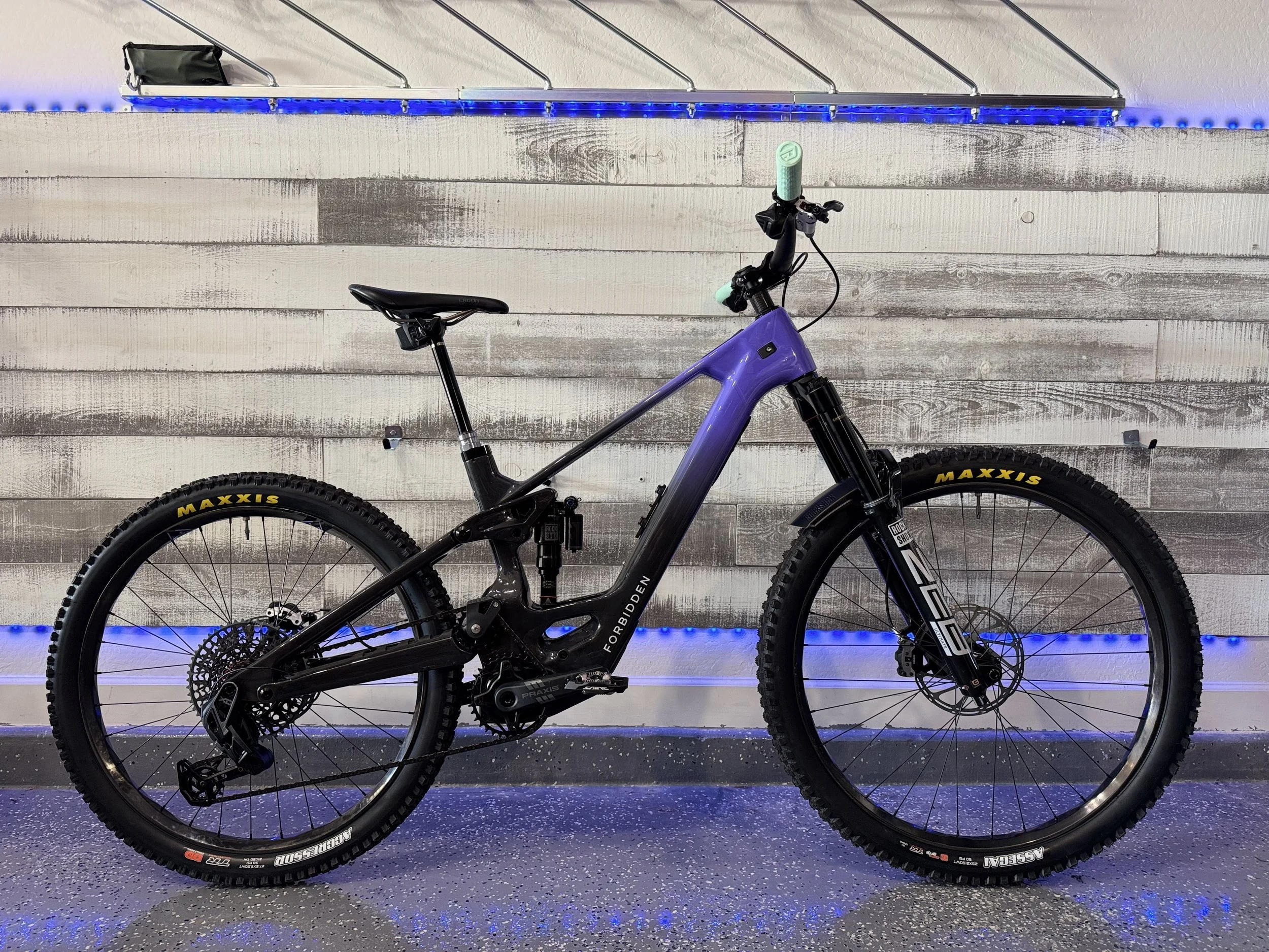 A purple full-suspension mountain bike with Maxxis tires is displayed against a white wooden wall with blue neon lighting. The bike features a sleek frame, disc brakes, and a saddle.