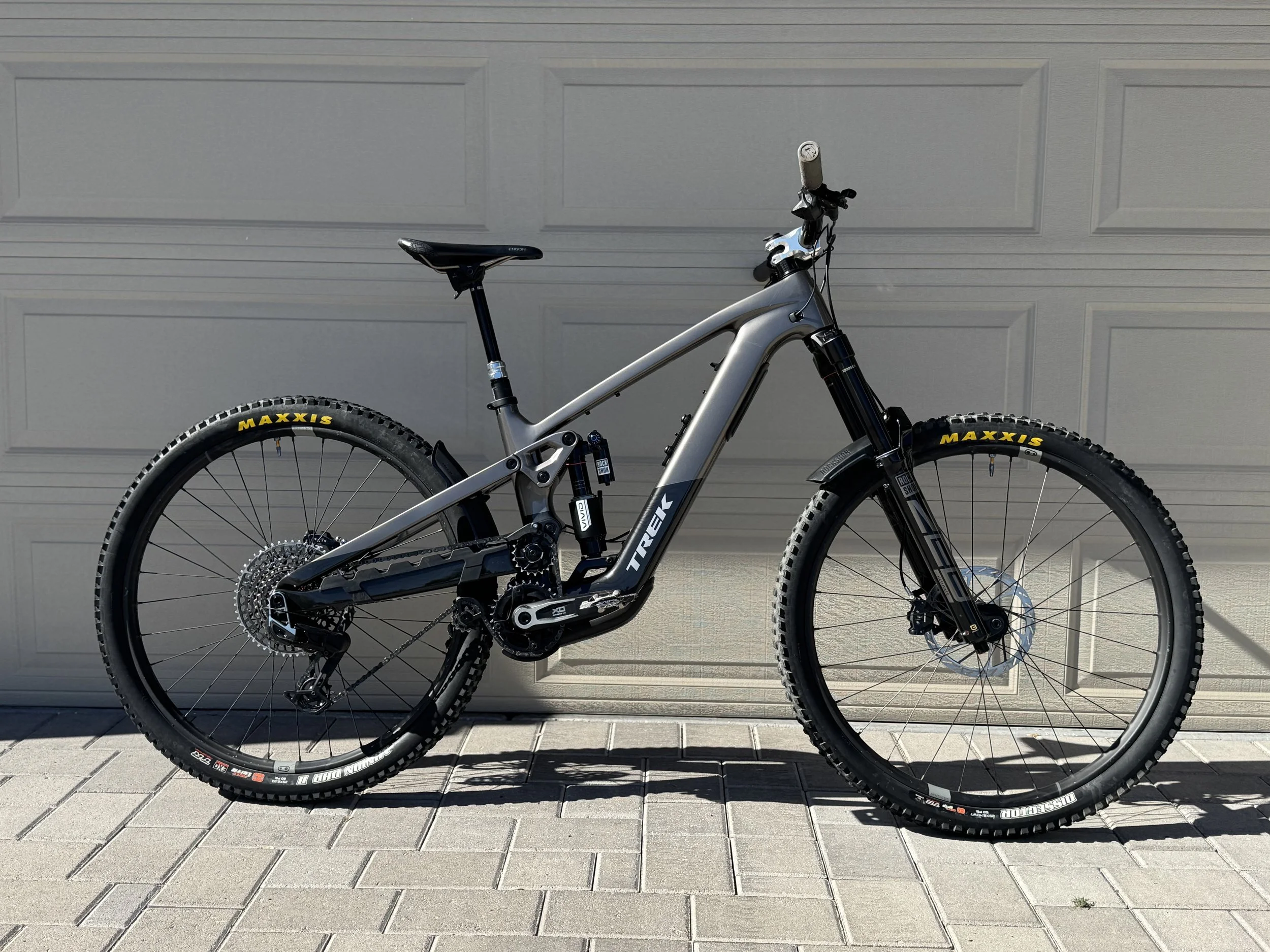 A full-suspension mountain bike with a silver frame, black front suspension fork, and Maxxis tires, parked against a gray garage door.