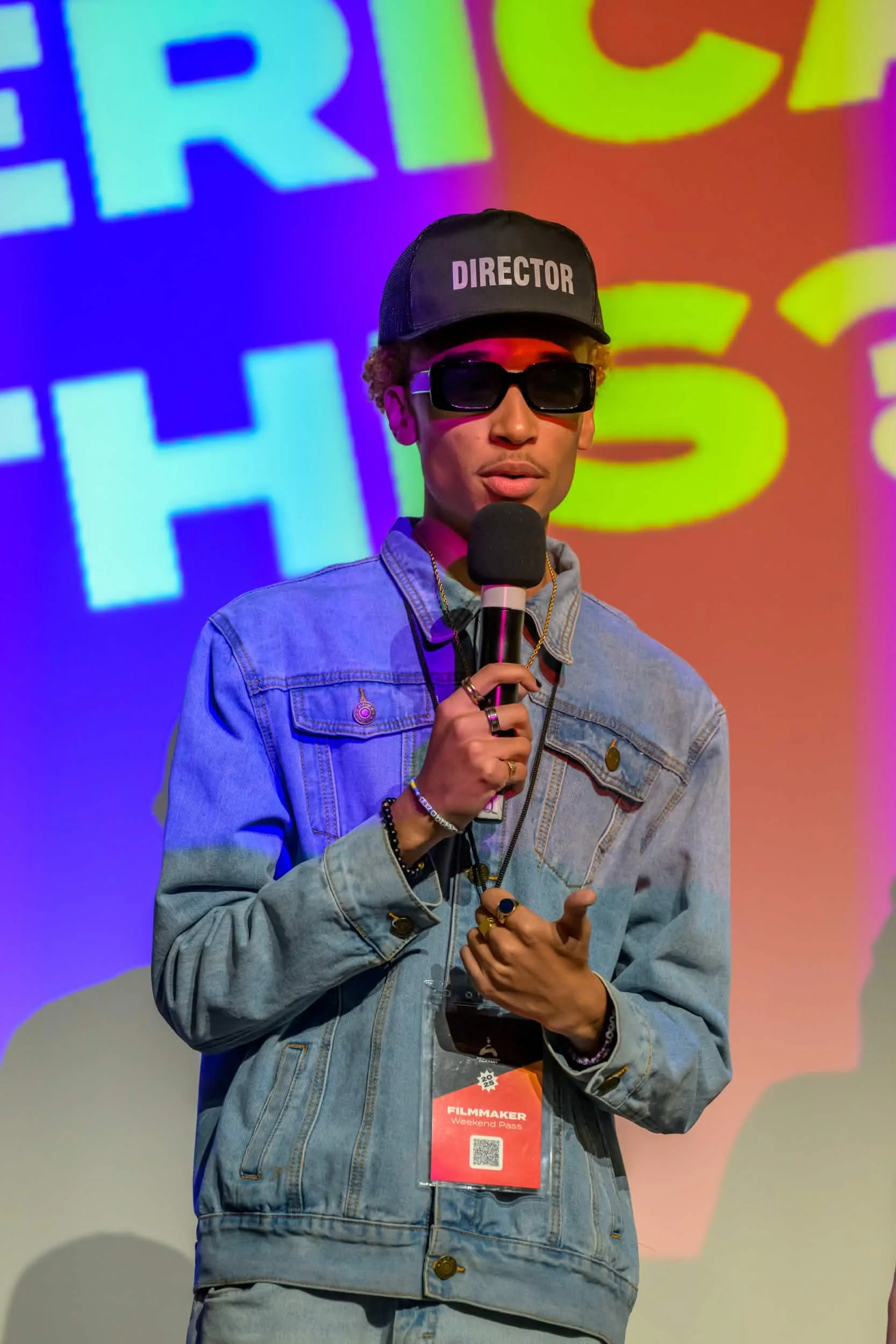 A young man wearing a black cap with the word 'DIRECTOR' in white letters, oversized black sunglasses, a denim jacket, and several rings, holding a microphone in his right hand, standing against a vibrant colorful background with large text.