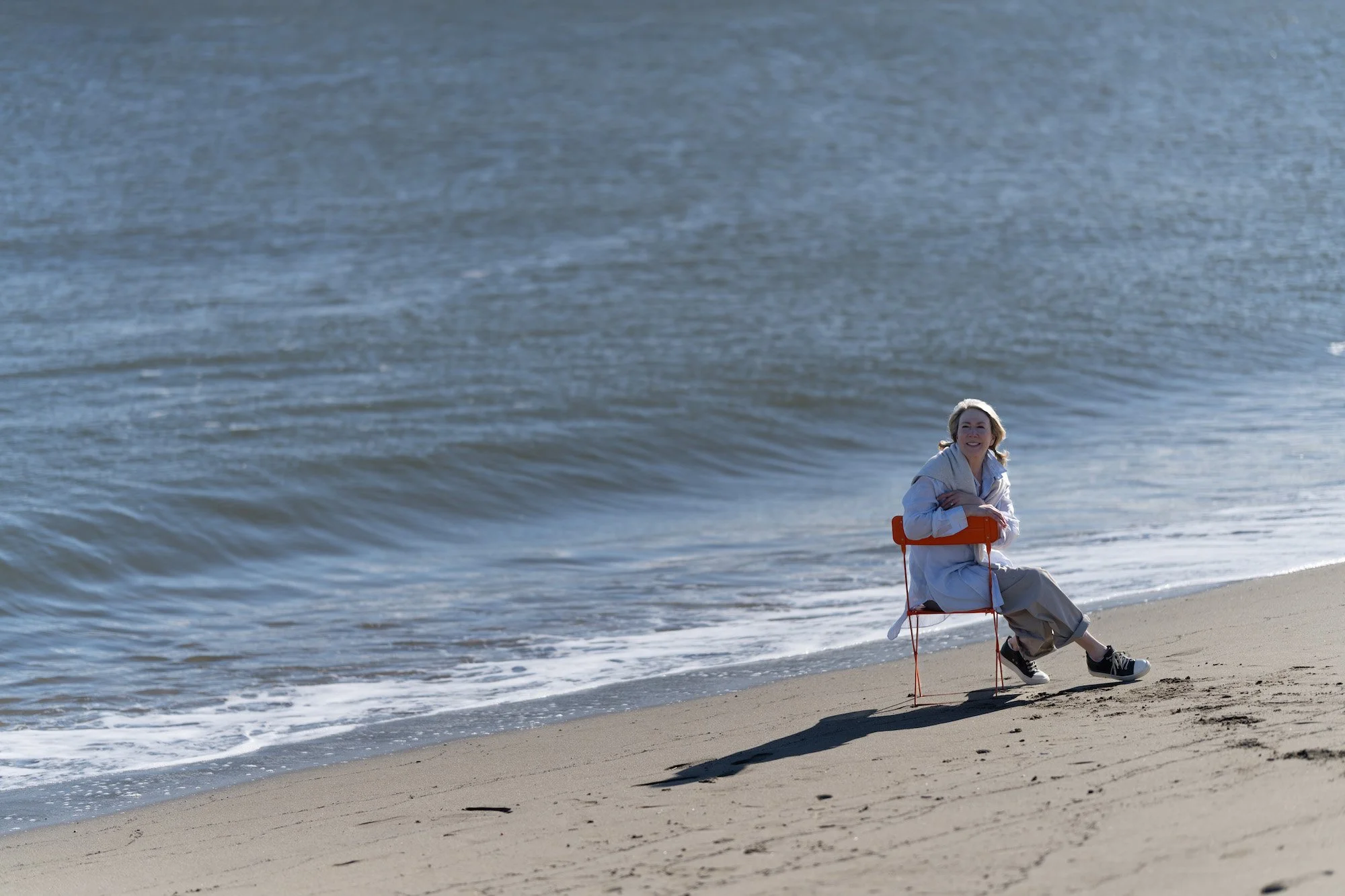 A woman sitting on a beach chair by the shoreline, smiling, with ocean waves in the background.