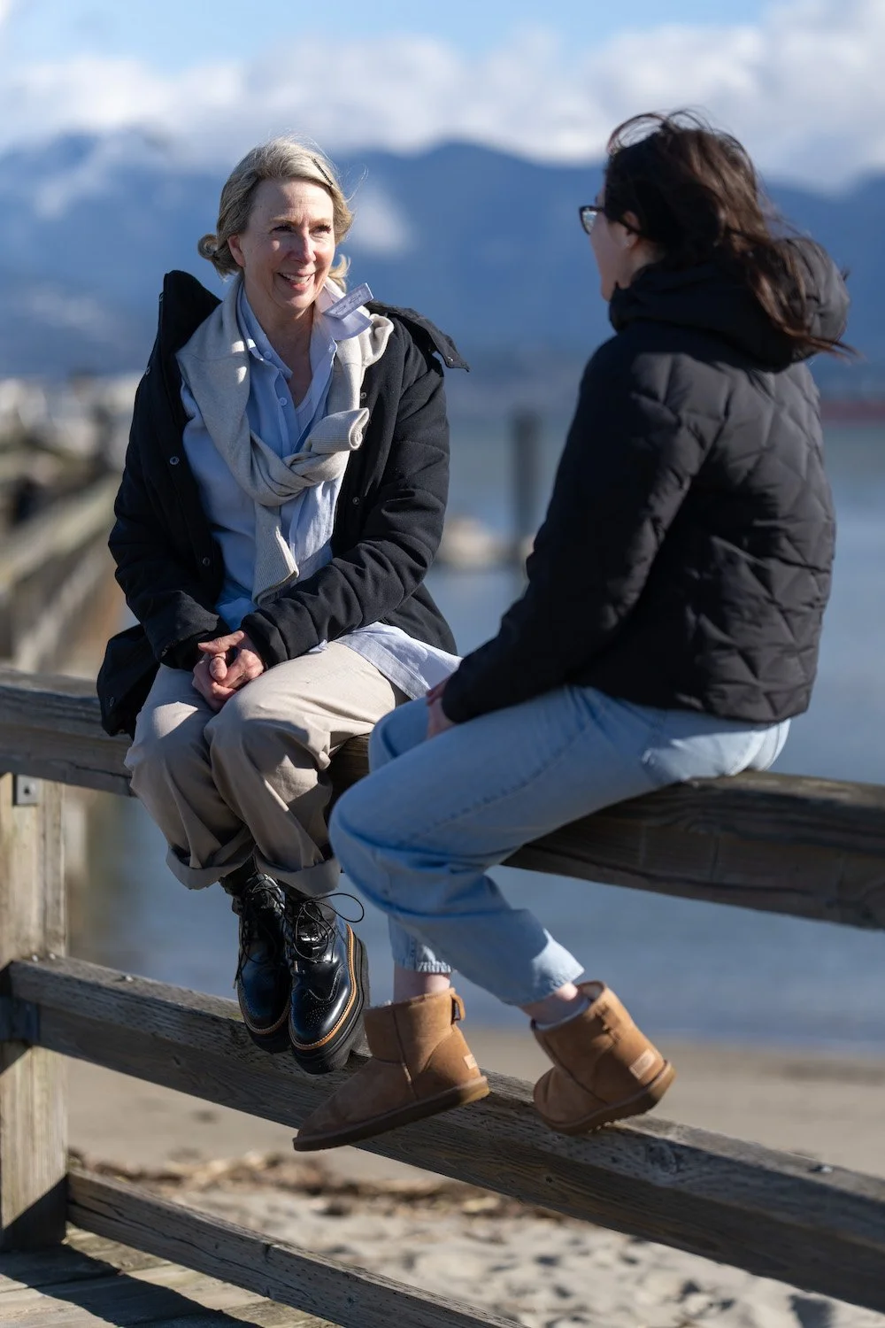 Two women sitting on a wooden railing by the water, engaged in conversation, with mountains and cloudy sky in the background.
