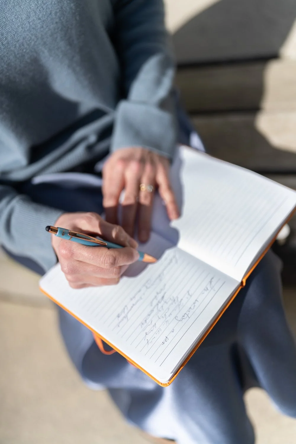 Person sitting outdoors writing in a notebook with a pen, wearing a gray sweater and a ring.