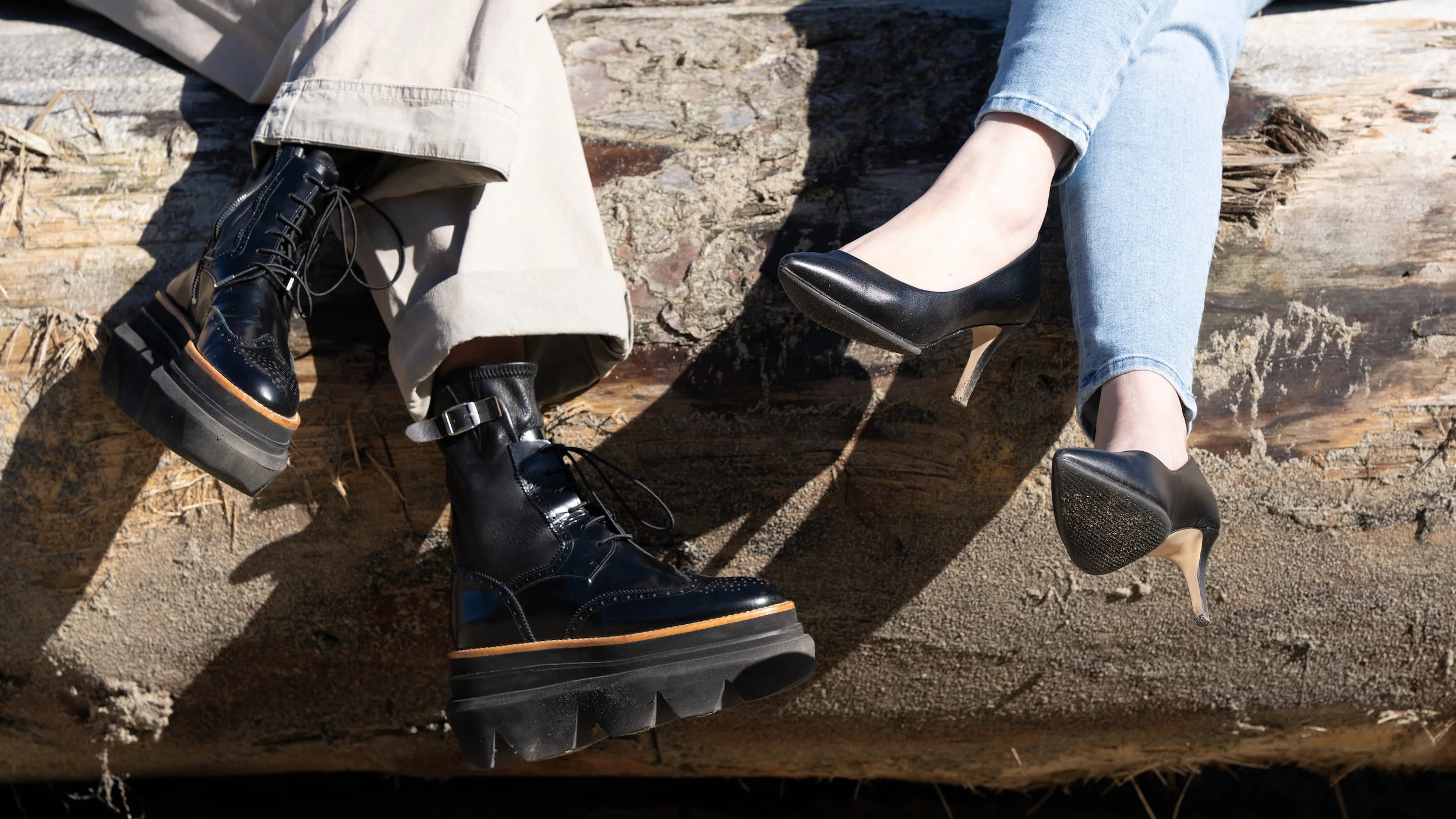A person's legs resting on a fallen tree branch, wearing light blue jeans and black high-heeled shoes, with another person's lower legs and feet visible, wearing beige pants and black chunky-sole combat boots.