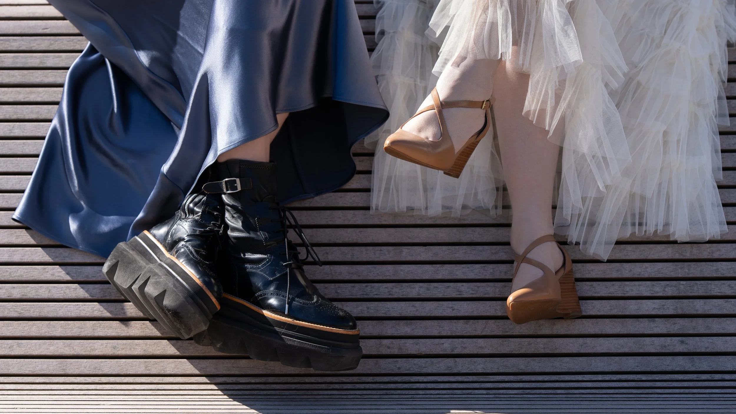 Close-up of two people sitting on a wooden deck, showing their legs and feet. One person wears black platform boots and a blue satin skirt, the other wears beige high-heeled shoes with a tulle skirt.