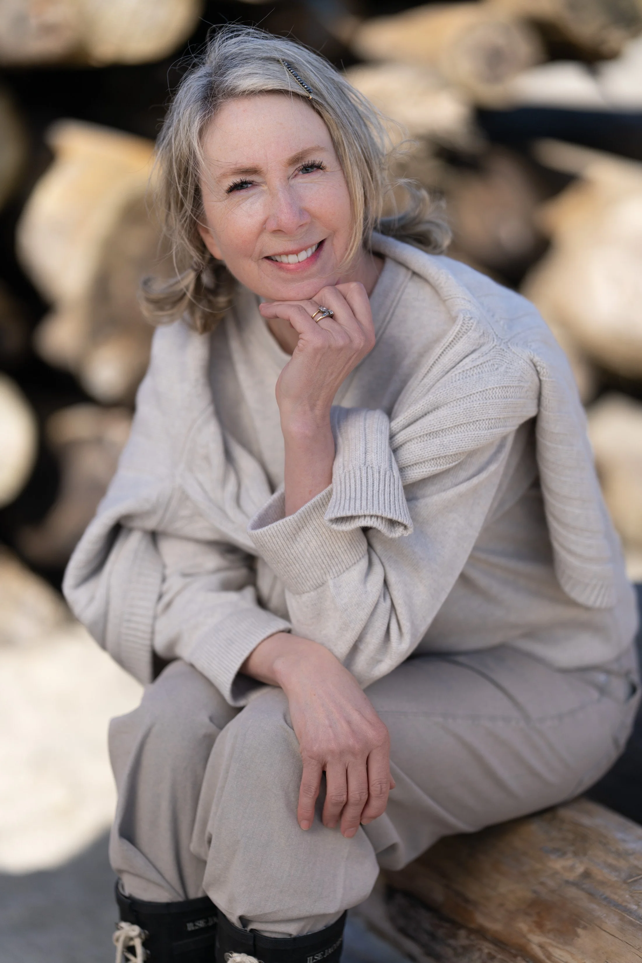 A woman with blonde hair smiling, wearing a light beige sweater, sitting on a wooden log, with a background of stacked logs.
