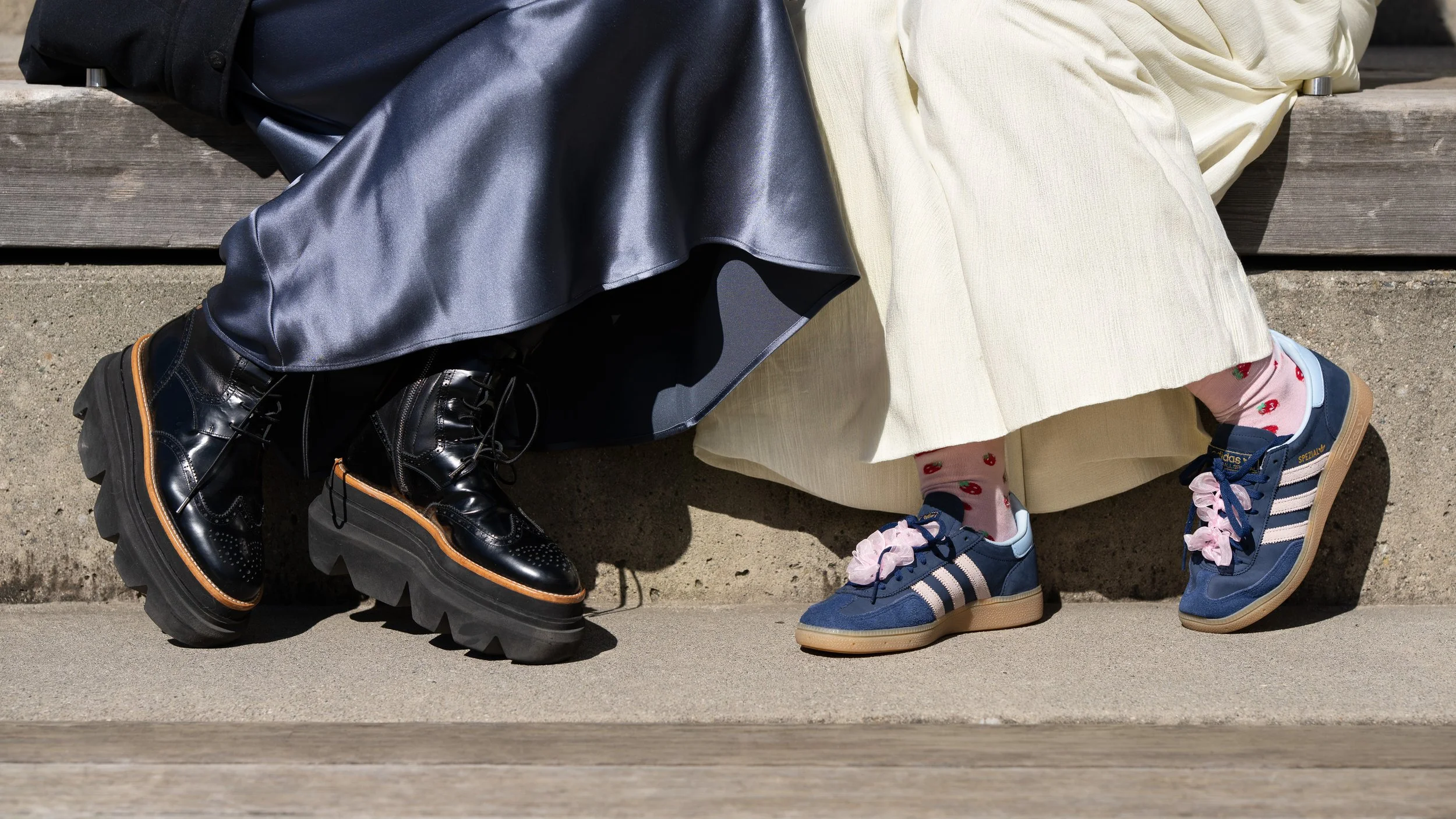 Close-up of two people sitting on a park bench, showing their legs and footwear. One person wears shiny black leather boots and a black skirt, the other wears colorful sneakers and a cream-colored dress.