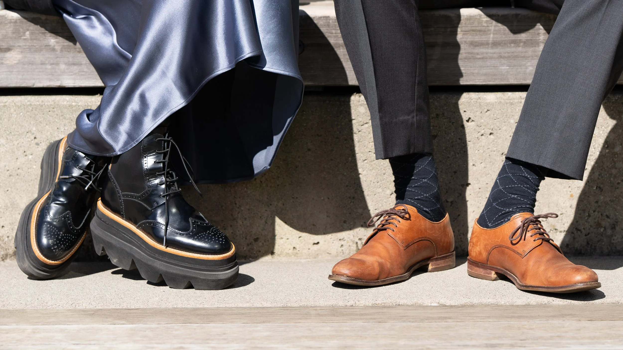 Close-up of two people wearing formal shoes, sitting on a bench. The person on the left has shiny black leather boots with thick soles, and the person on the right has tan leather dress shoes. Both are wearing dark trousers and patterned socks.