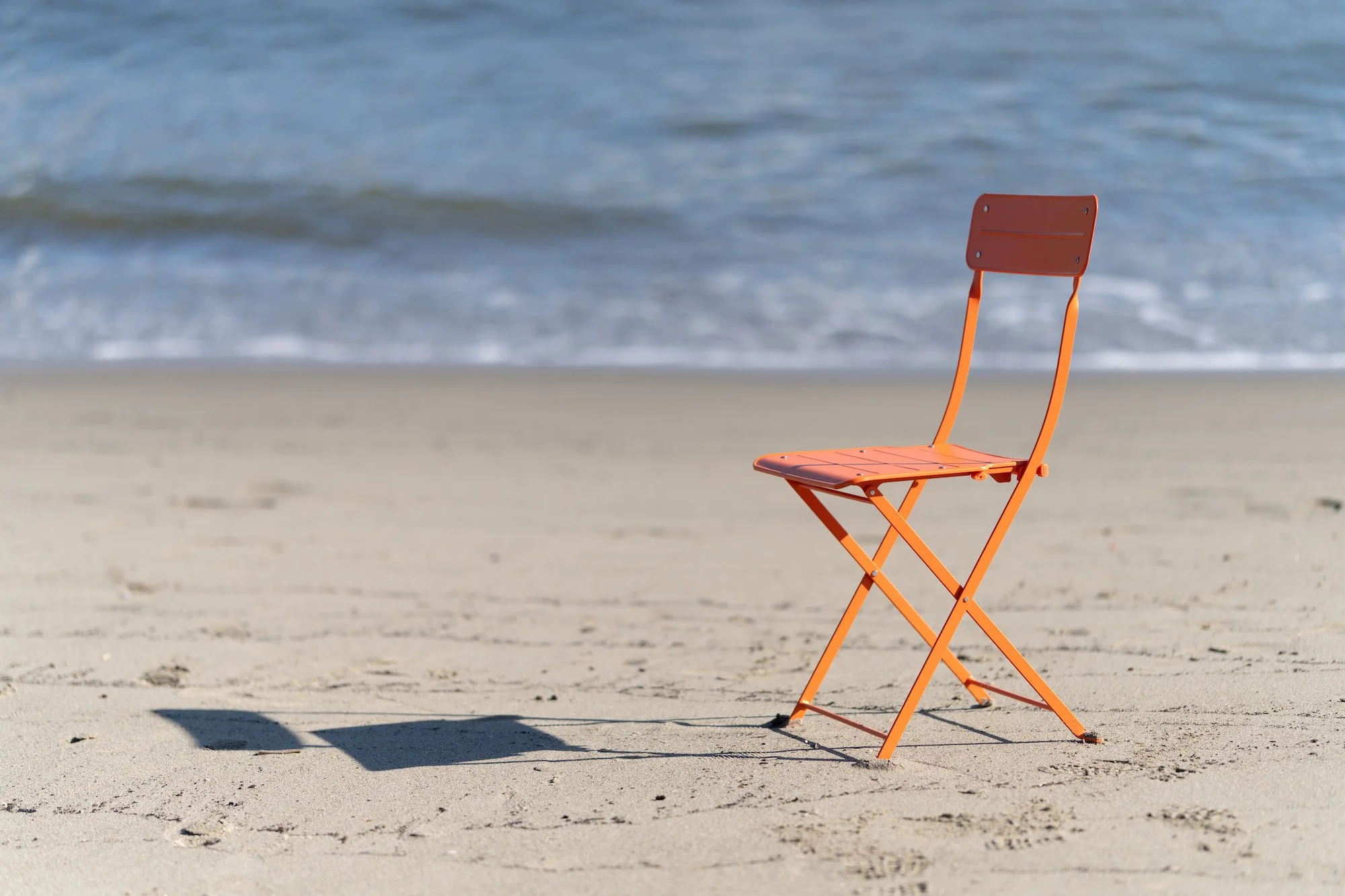 An orange folding chair on a sandy beach near the ocean, with waves in the background.