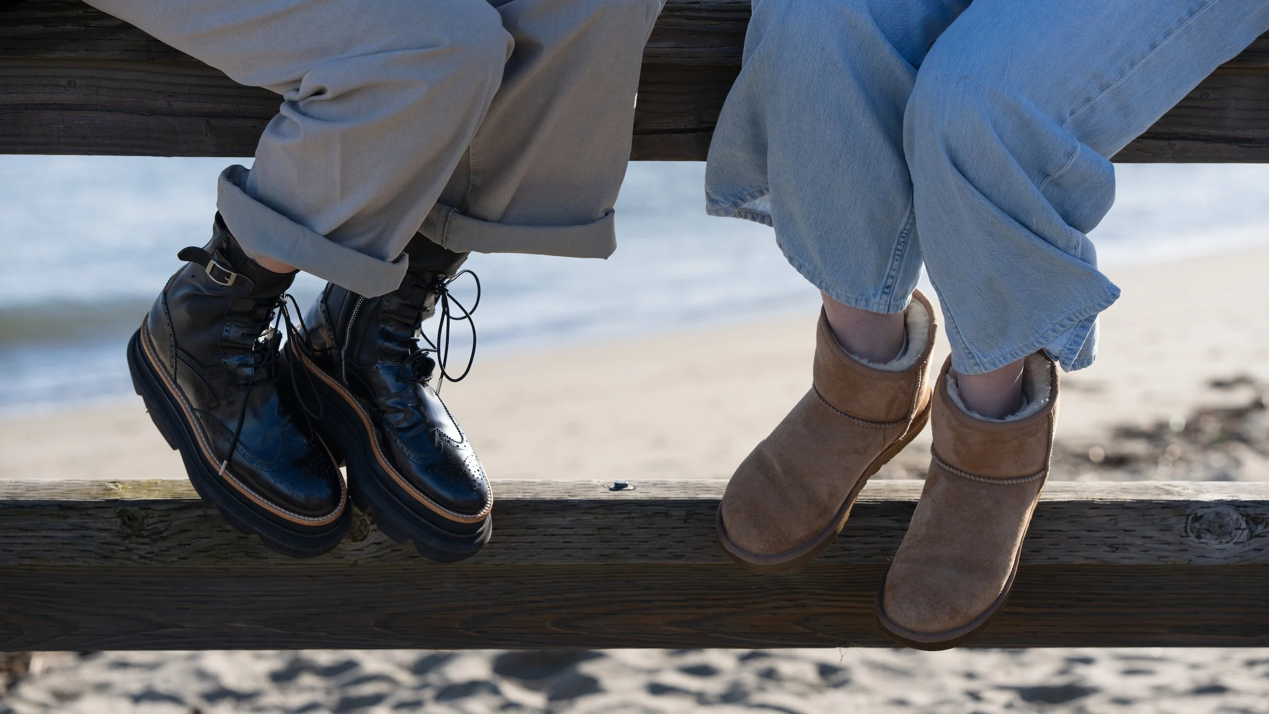 People sitting on a wooden beam at the beach, wearing boots with the ocean in the background.