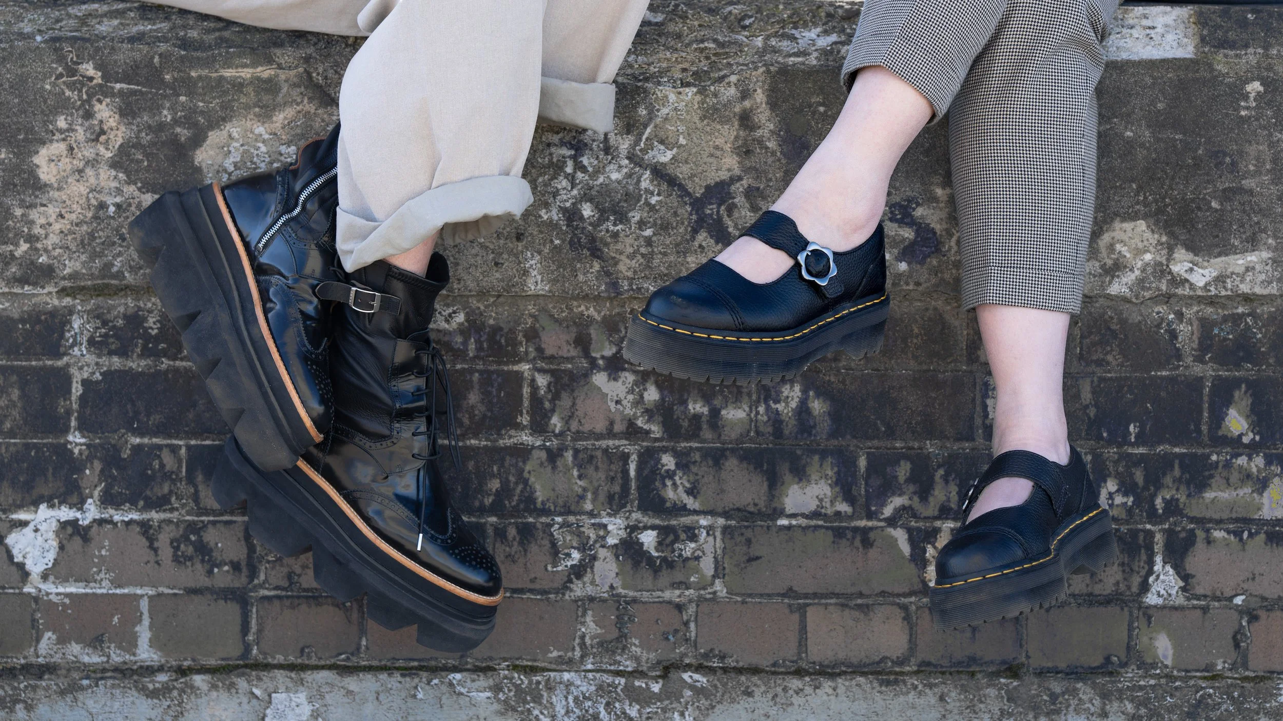 Close-up of two people sitting on a brick ledge, showing their legs and feet, both wearing black platform shoes with thick soles, one with an ankle strap, against a graffiti-covered wall.