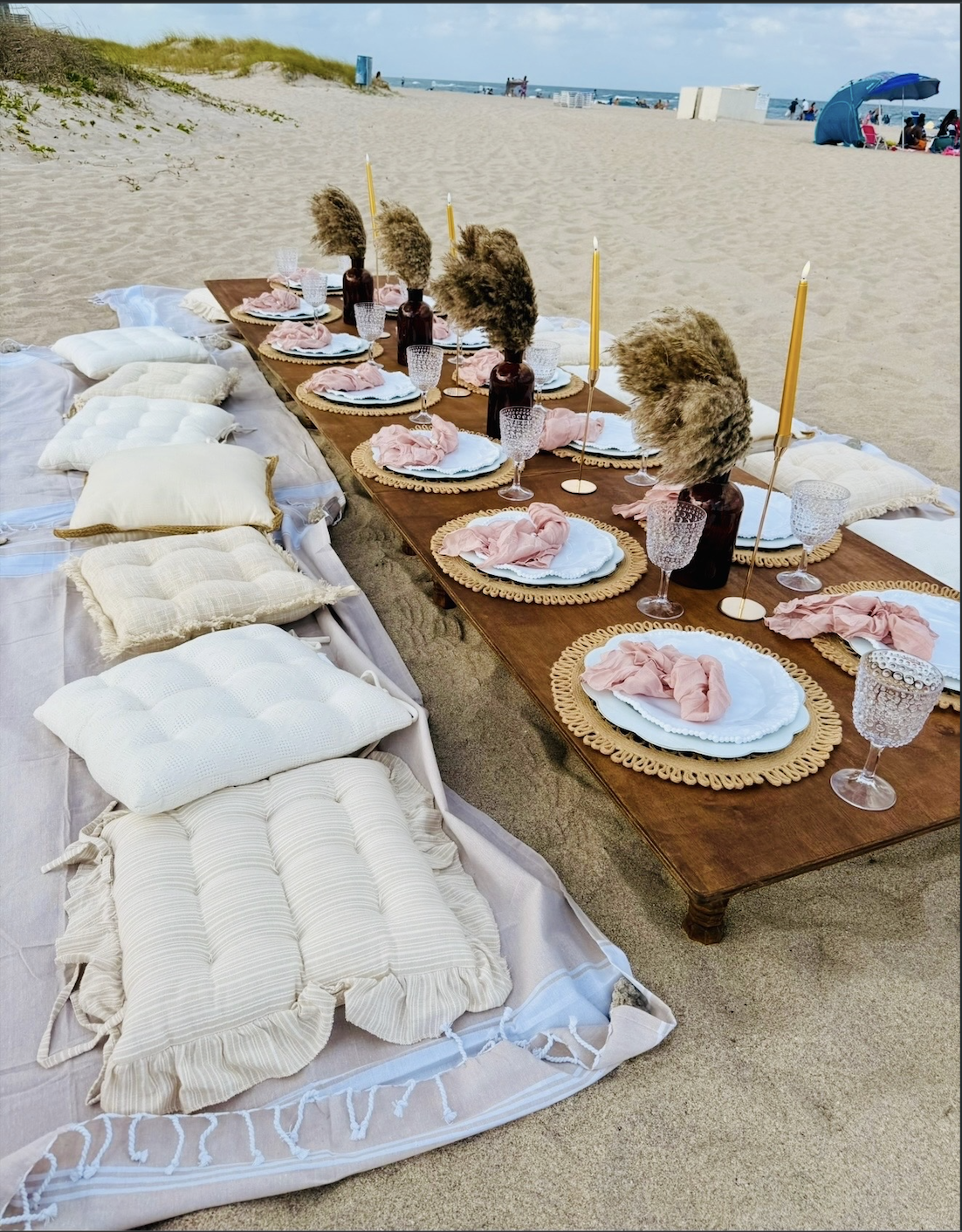 A beach picnic setup with a long wooden table arranged on the sand, decorated with pink napkins, white plates, pink and white place mats, and glassware. Cushions and blankets are laid out on the sand beside the table for seating. The background shows