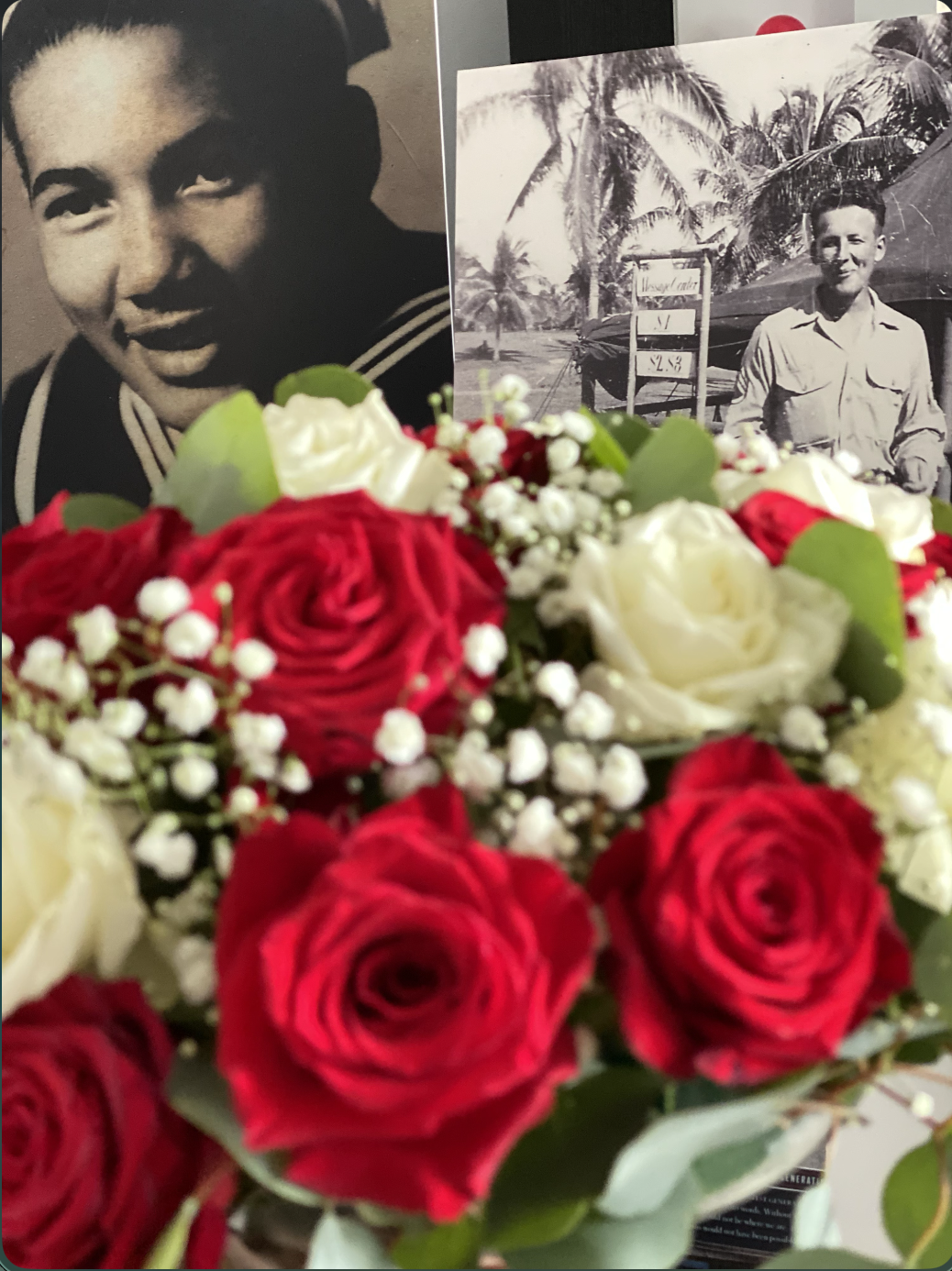 A bouquet of red and white roses with white baby's breath flowers and green leaves in the foreground. Behind the bouquet are two black-and-white photos of a young man, one in uniform and the other outdoors with palm trees and a sign showing prices.