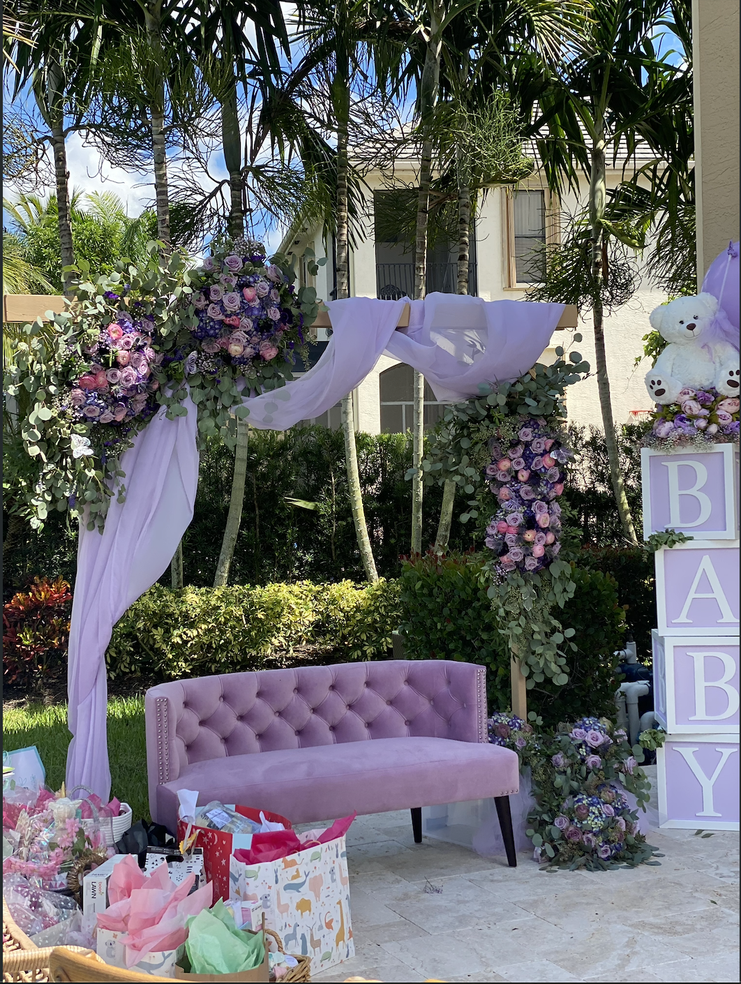 Decorative baby shower setup with pink tufted loveseat, floral arch, purple and pink flowers, white teddy bear, and baby blocks spelling 'BABY' outdoors with trees and house in background.