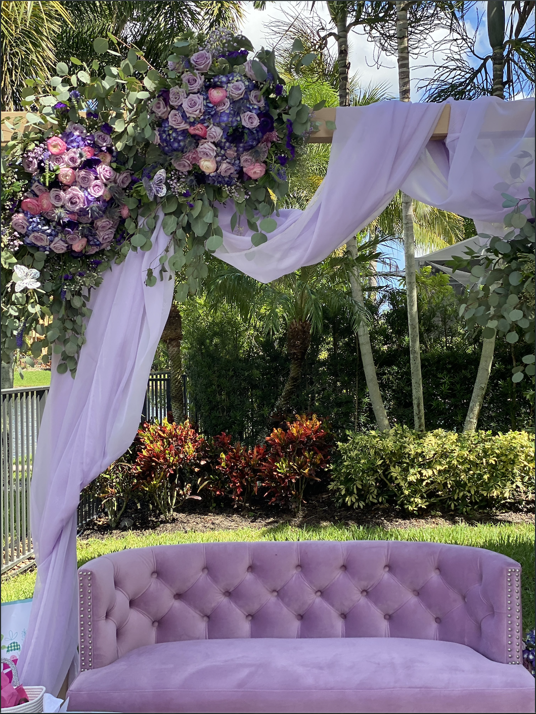A pink tufted velvet sofa beneath an arch decorated with purple and pink flowers and white drapery, set outdoors with green trees and plants in the background.