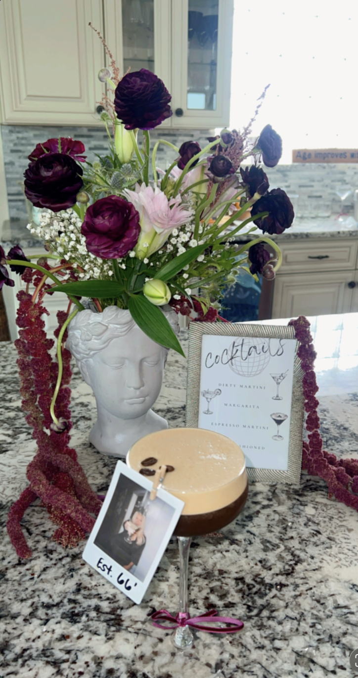 A decorative countertop display featuring a white bust-shaped vase filled with a colorful flower arrangement including purple and pink blooms with greenery, a framed cocktail menu, a photograph clipped to a string, and a creamy coffee-colored cocktai