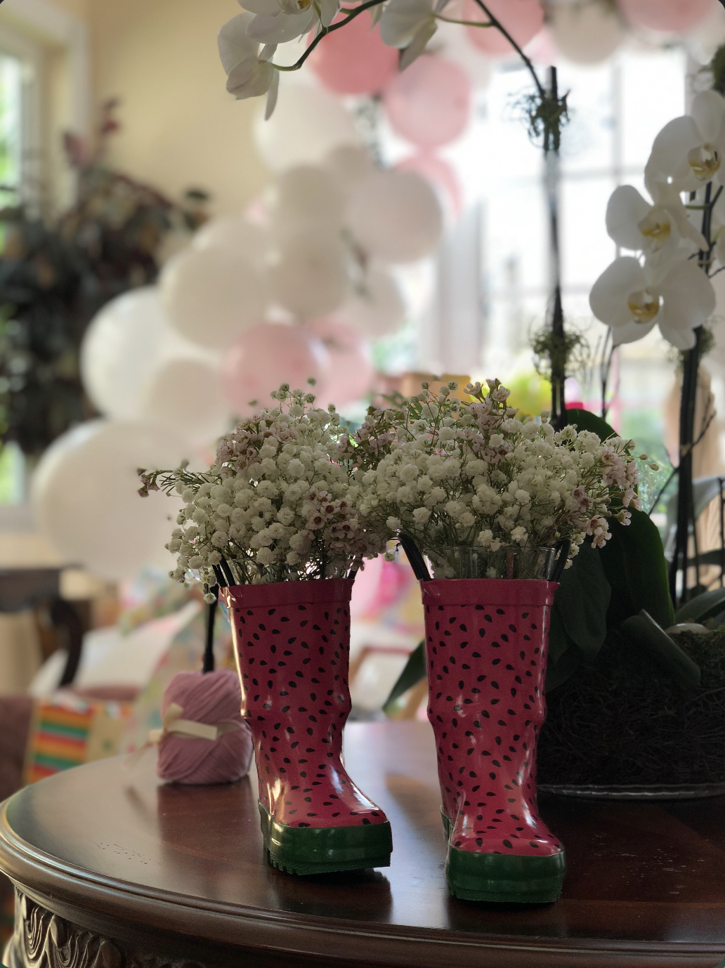 Pink rain boots repurposed as flower vases filled with white and pink flowers, on a wooden table with balloons and other decorations in the background.