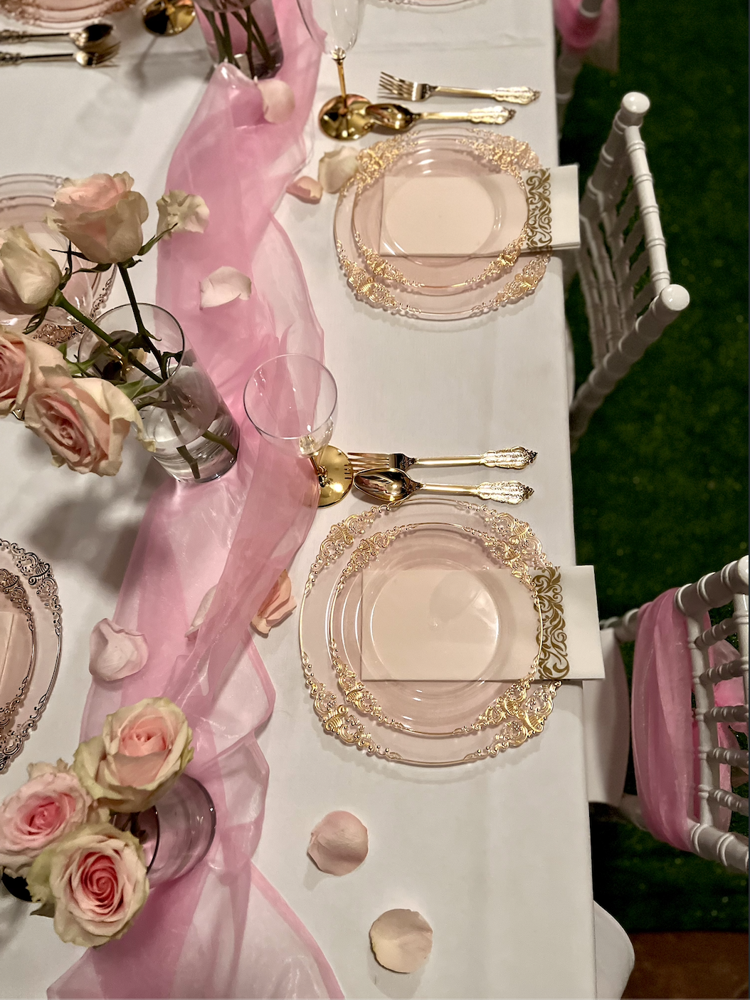 Elegant table setting with pink and gold accents, featuring clear glass plates with gold trim, gold utensils, pink cloth napkins, and a centerpiece of pink and white roses in glass vases on a white tablecloth with pink sheer fabric and scattered rose