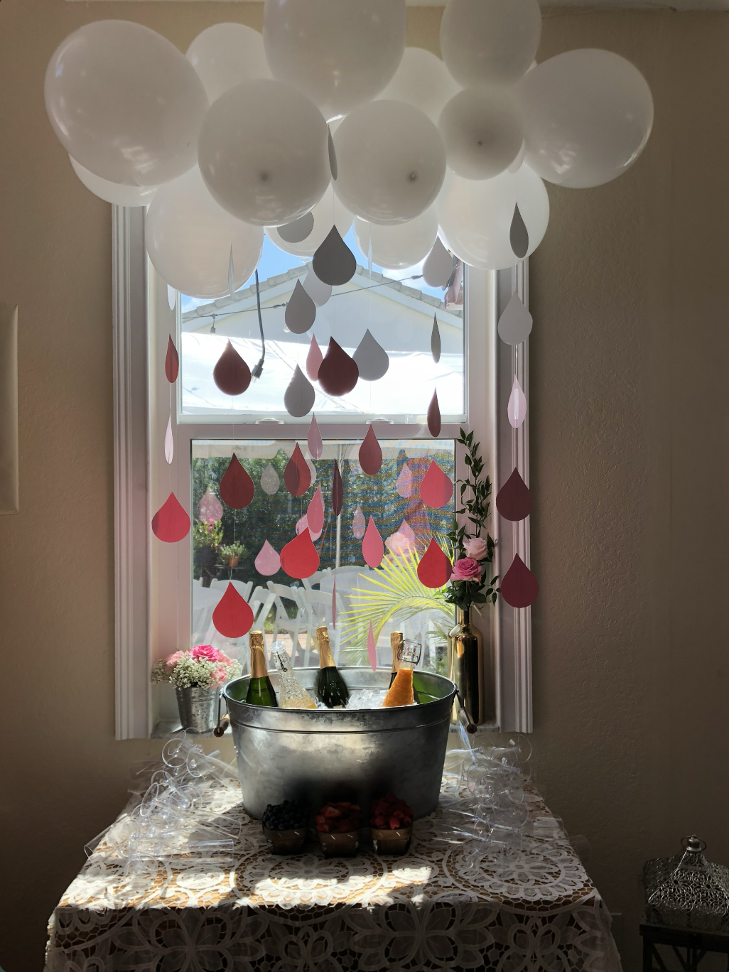 Decorative indoor setup with white and pink hanging paper raindrop cutouts from a cluster of white balloons, a window with outside view, a table covered with a lace tablecloth holding a metallic bucket with bottles of champagne and sparkling wine, su